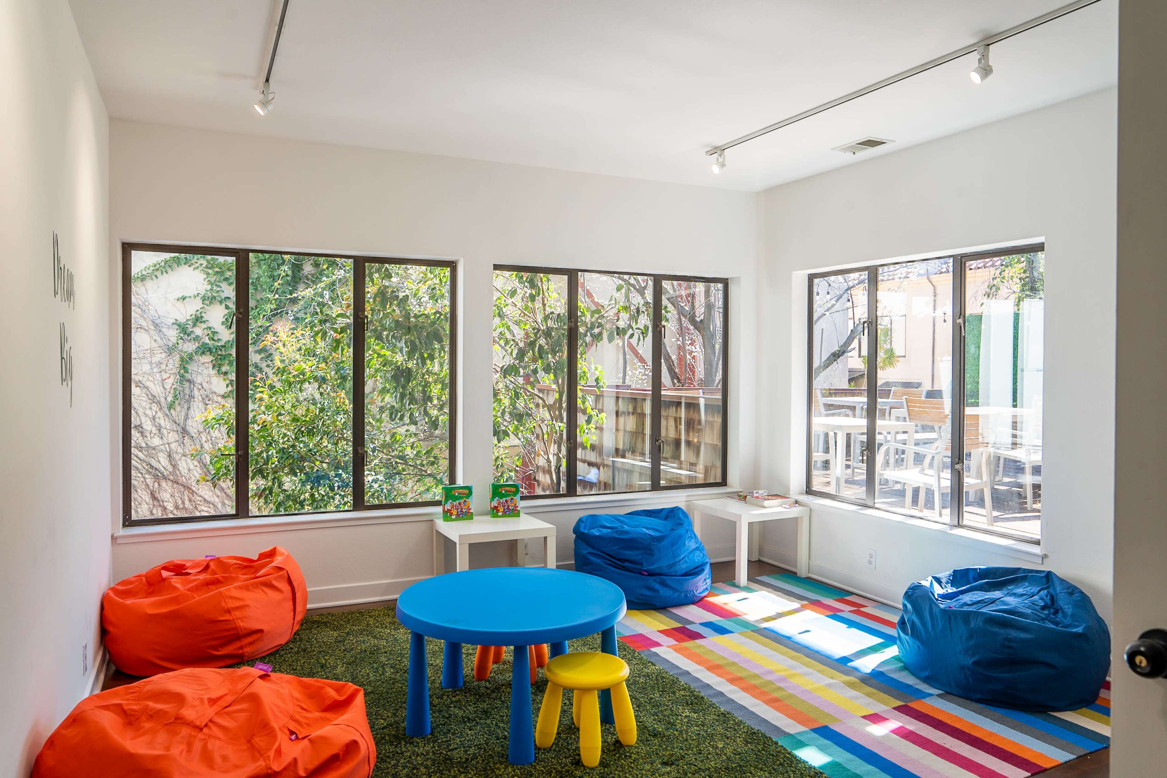 A playroom features colorful bean bags, a round blue table, and large windows that provide natural light and a view of greenery outside.
