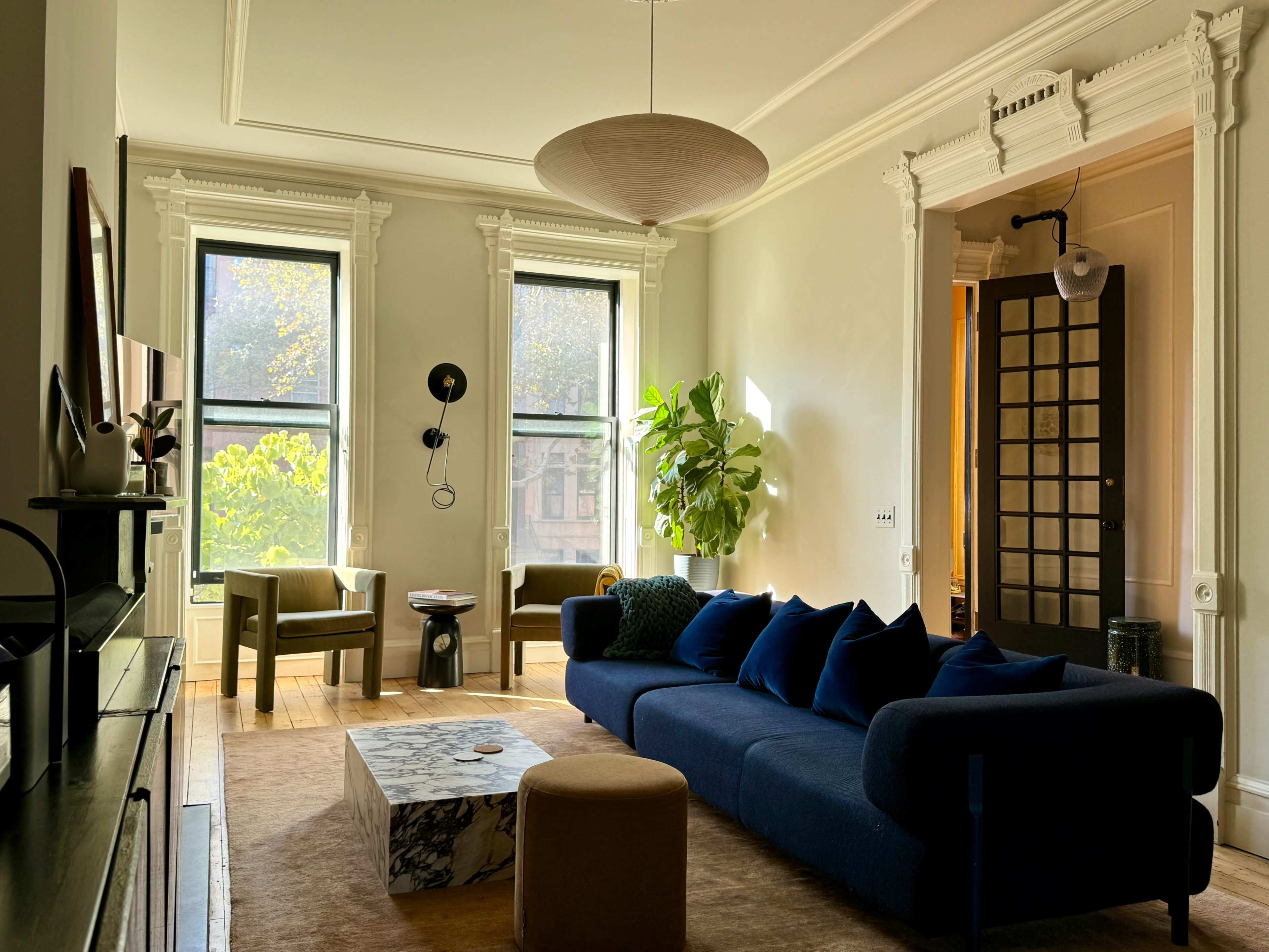 The room features a modern sofa, a marble-topped coffee table, two armchairs, and large windows with a view of greenery.