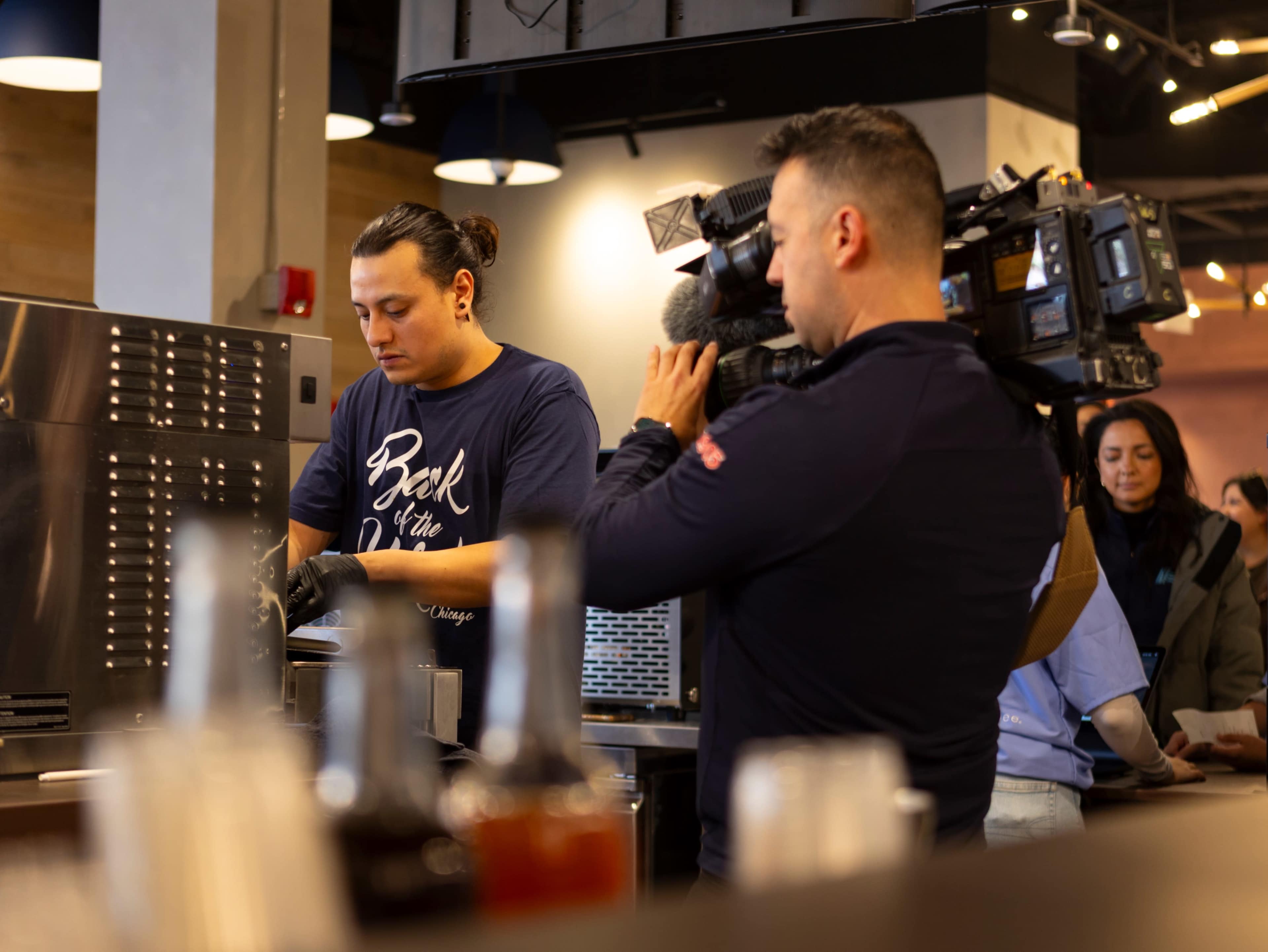 A cook prepares food in a restaurant kitchen while a cameraman films the activity.