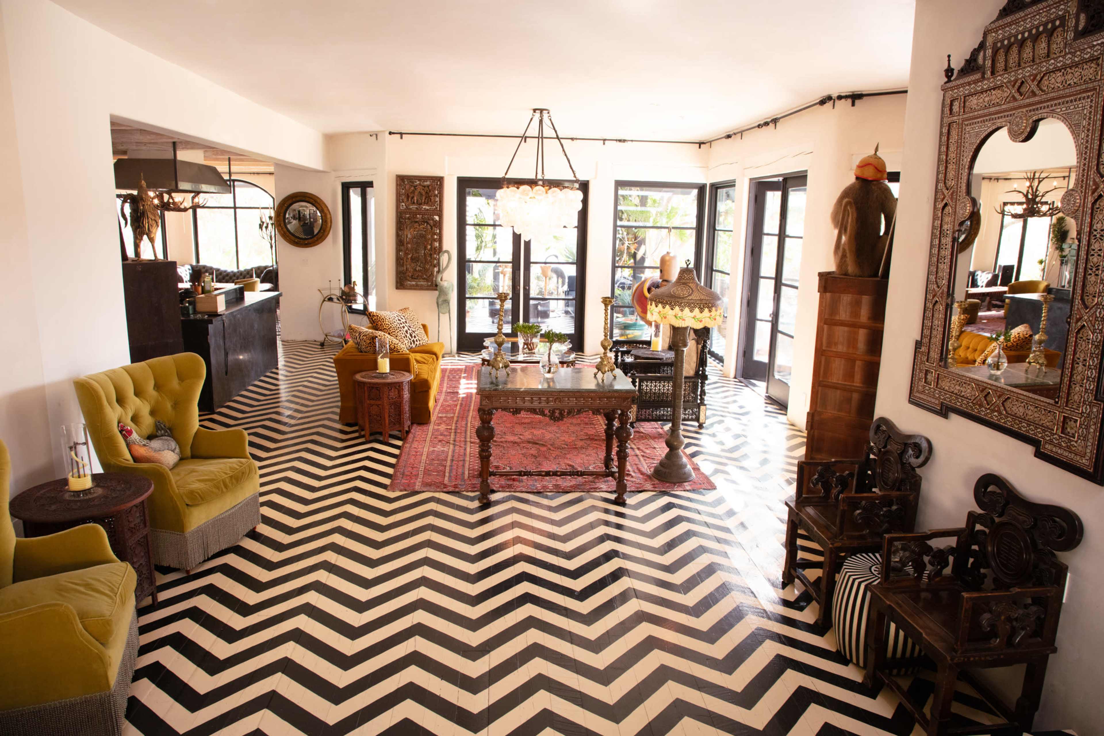 The image shows a spacious living room with a chevron-patterned floor, eclectic furniture, and large windows allowing natural light to enter.