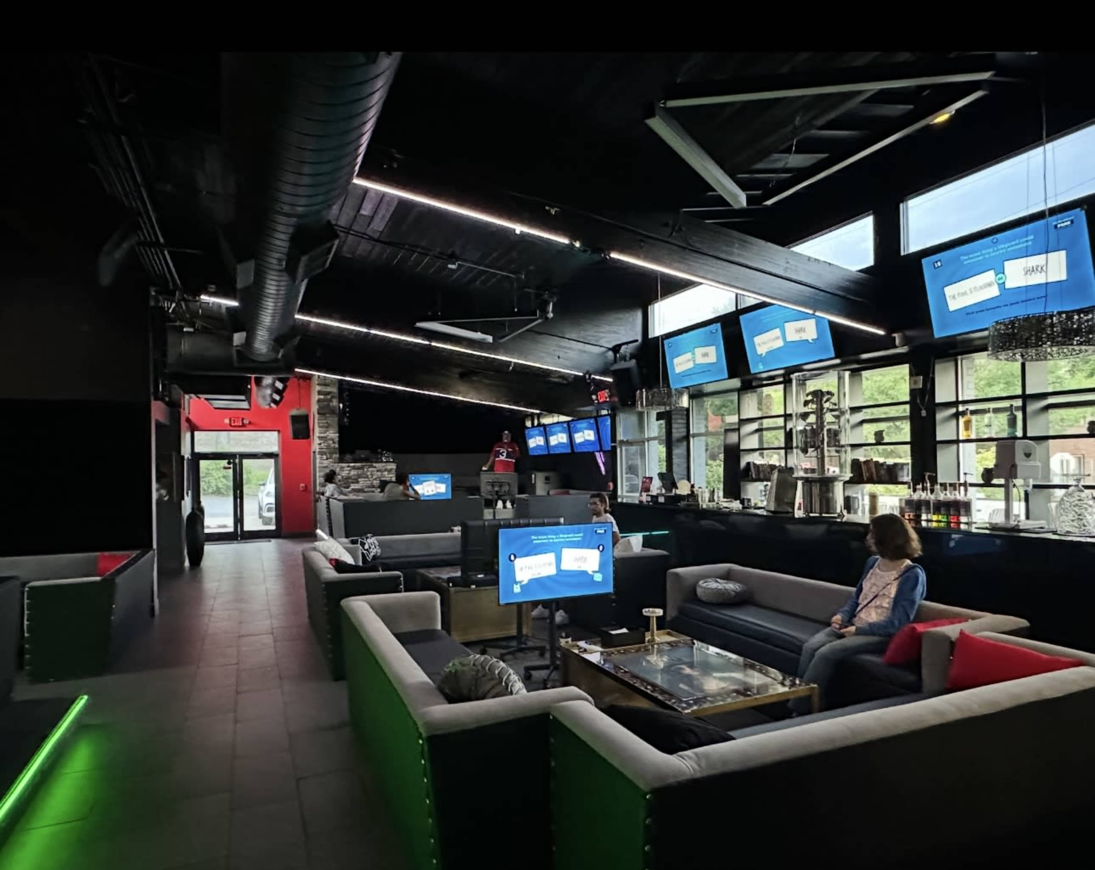 The image shows a modern lounge area with several seating arrangements, large screens displaying content, and a bar section in the background.