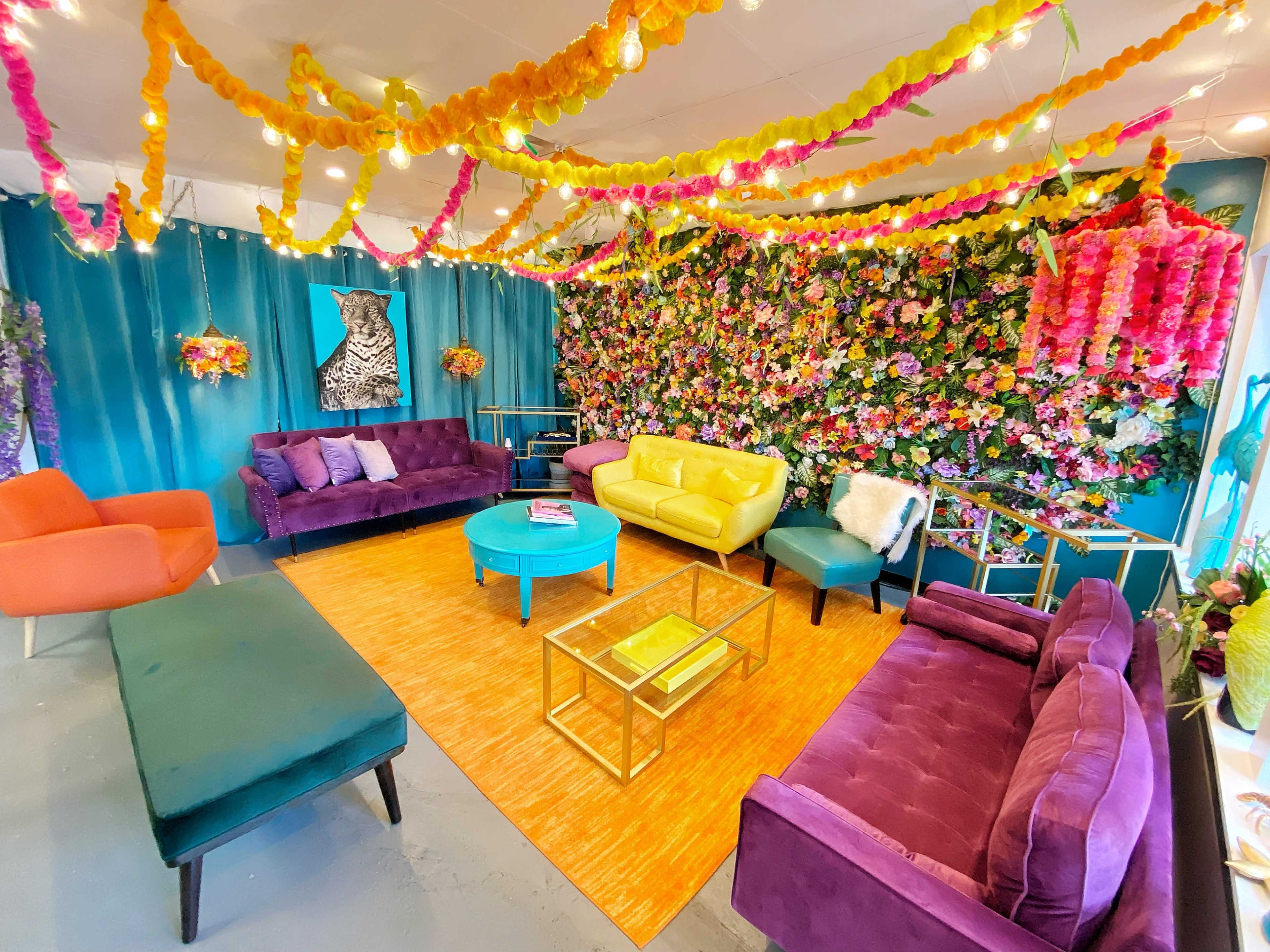The room features colorful furniture arranged around a vibrant floral wall, with decorative streamers hanging from the ceiling.