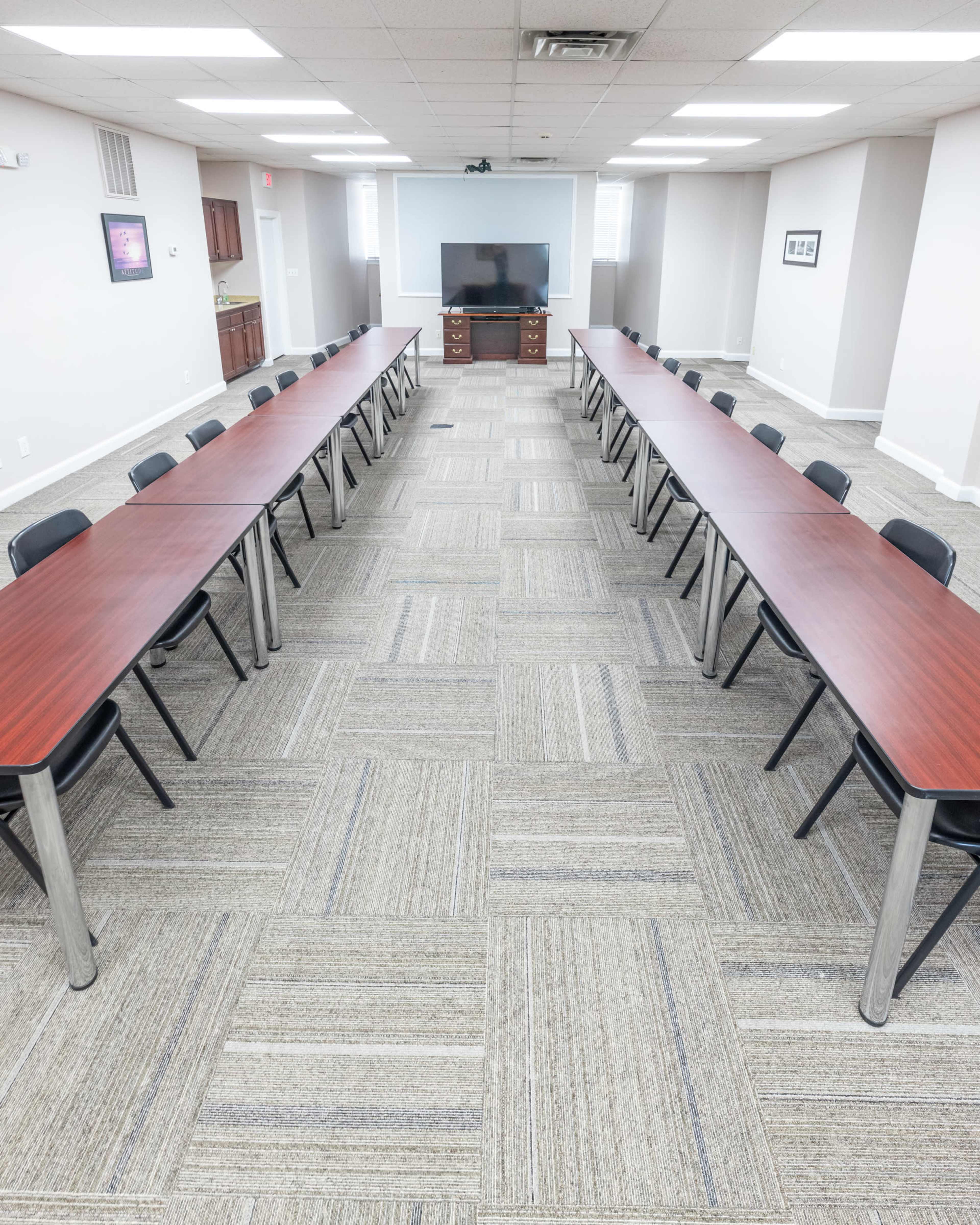 A conference room features two long tables arranged in a U-shape with black chairs, and a large screen mounted on the wall.