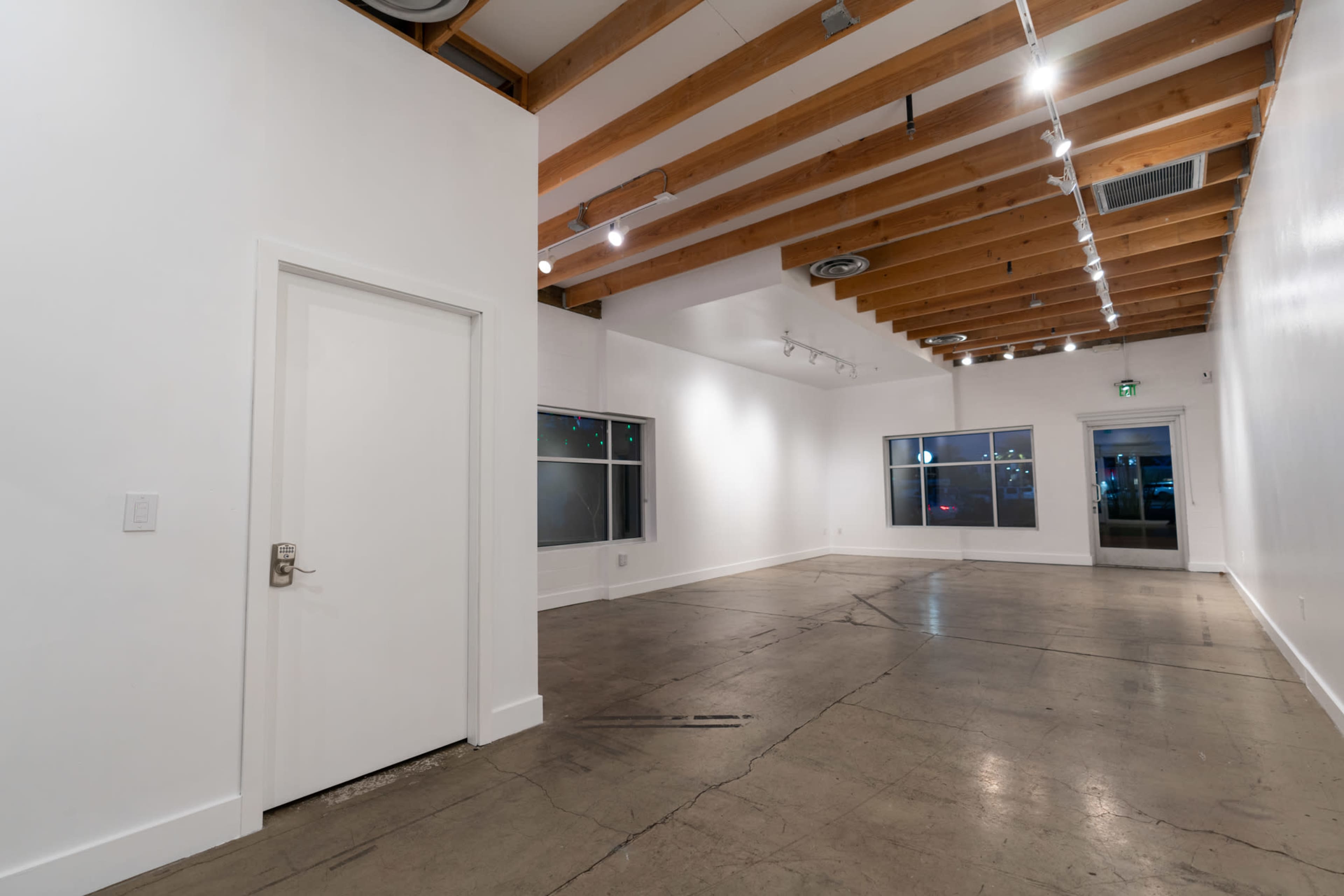 An empty room with white walls, exposed wooden beams, concrete floors, and large windows.