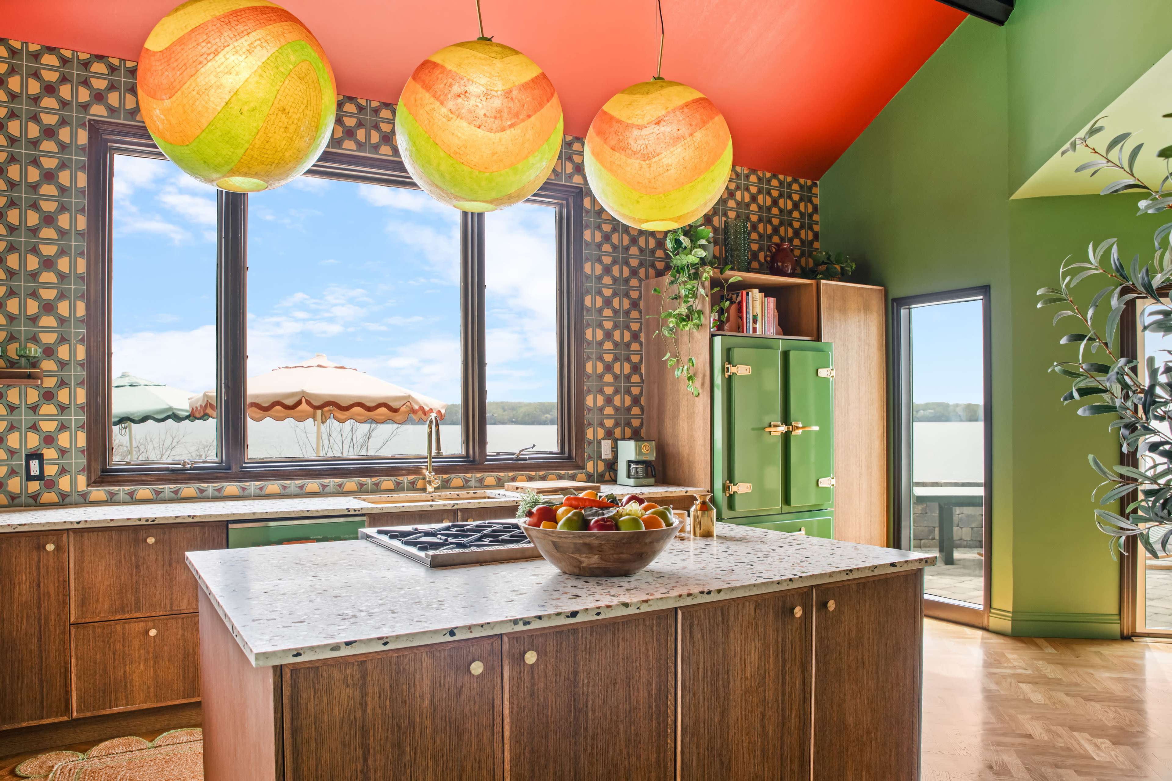 The kitchen features a central island, colorful pendant lights, and a patterned backsplash, with large windows offering a view of an outdoor patio.