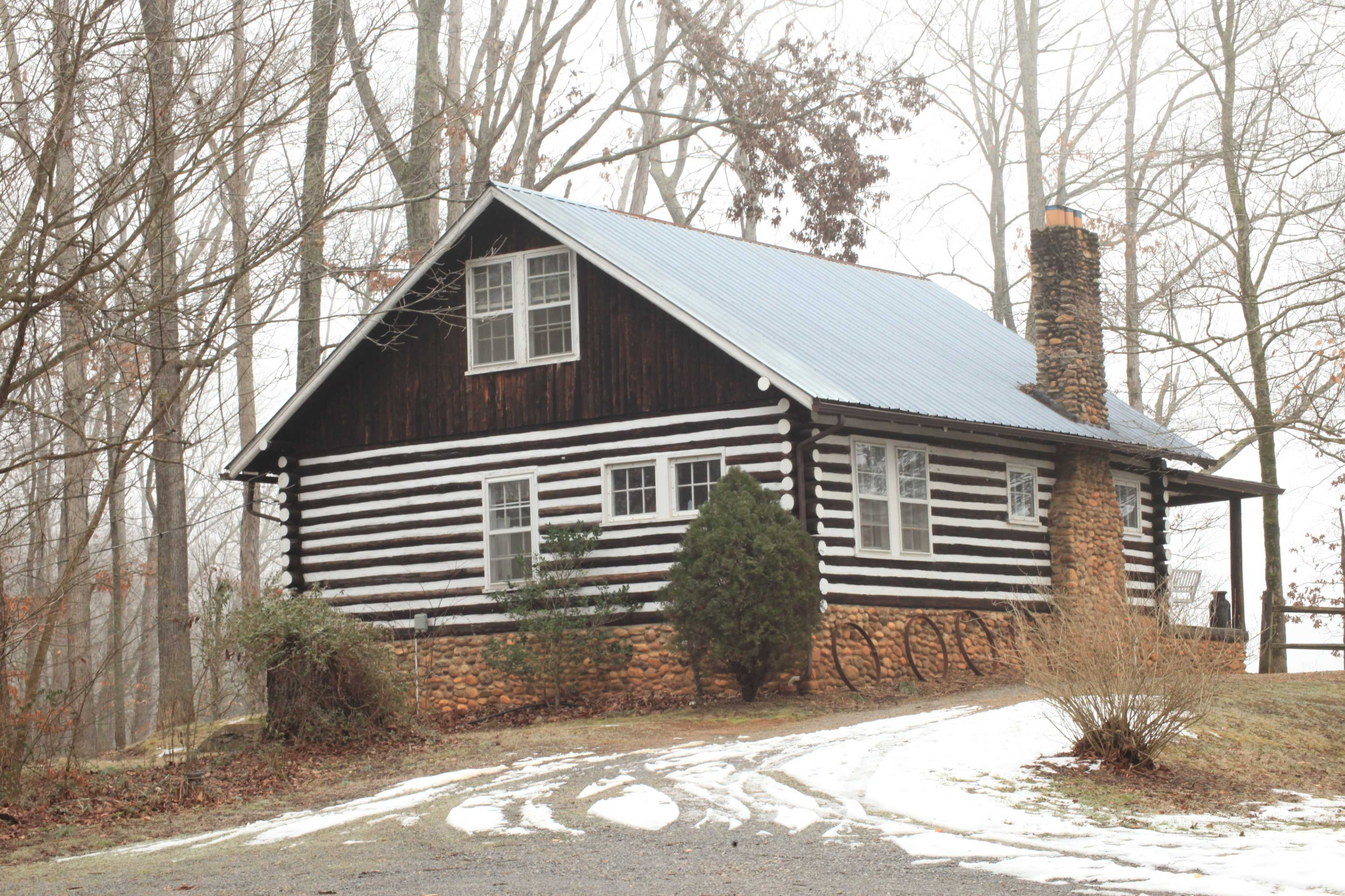 A log cabin with a stone chimney sits on a gravel path surrounded by bare trees and patches of snow.