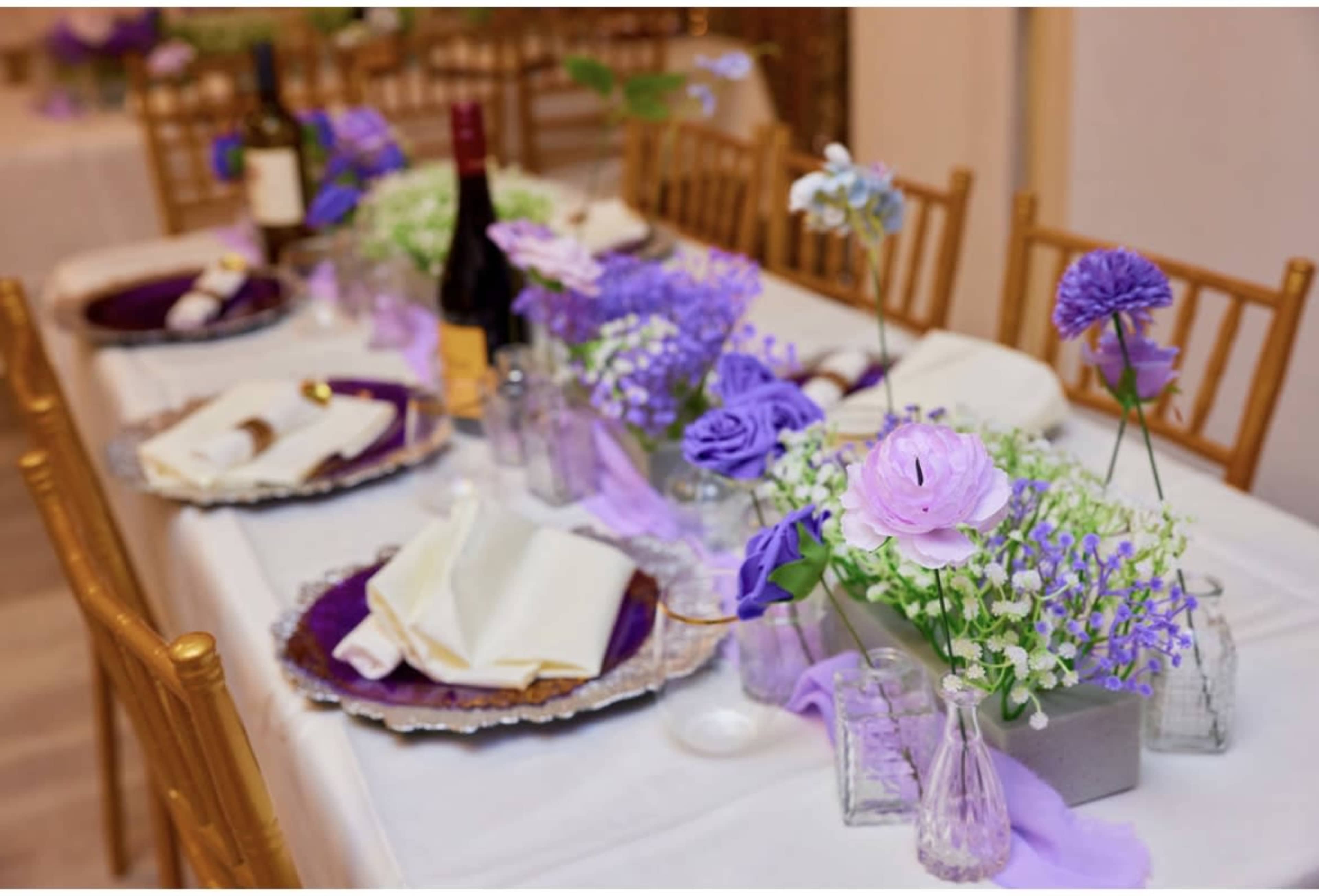 The scene features a beautifully arranged dining table set with purple floral centerpieces, wine bottles, and elegant tableware.