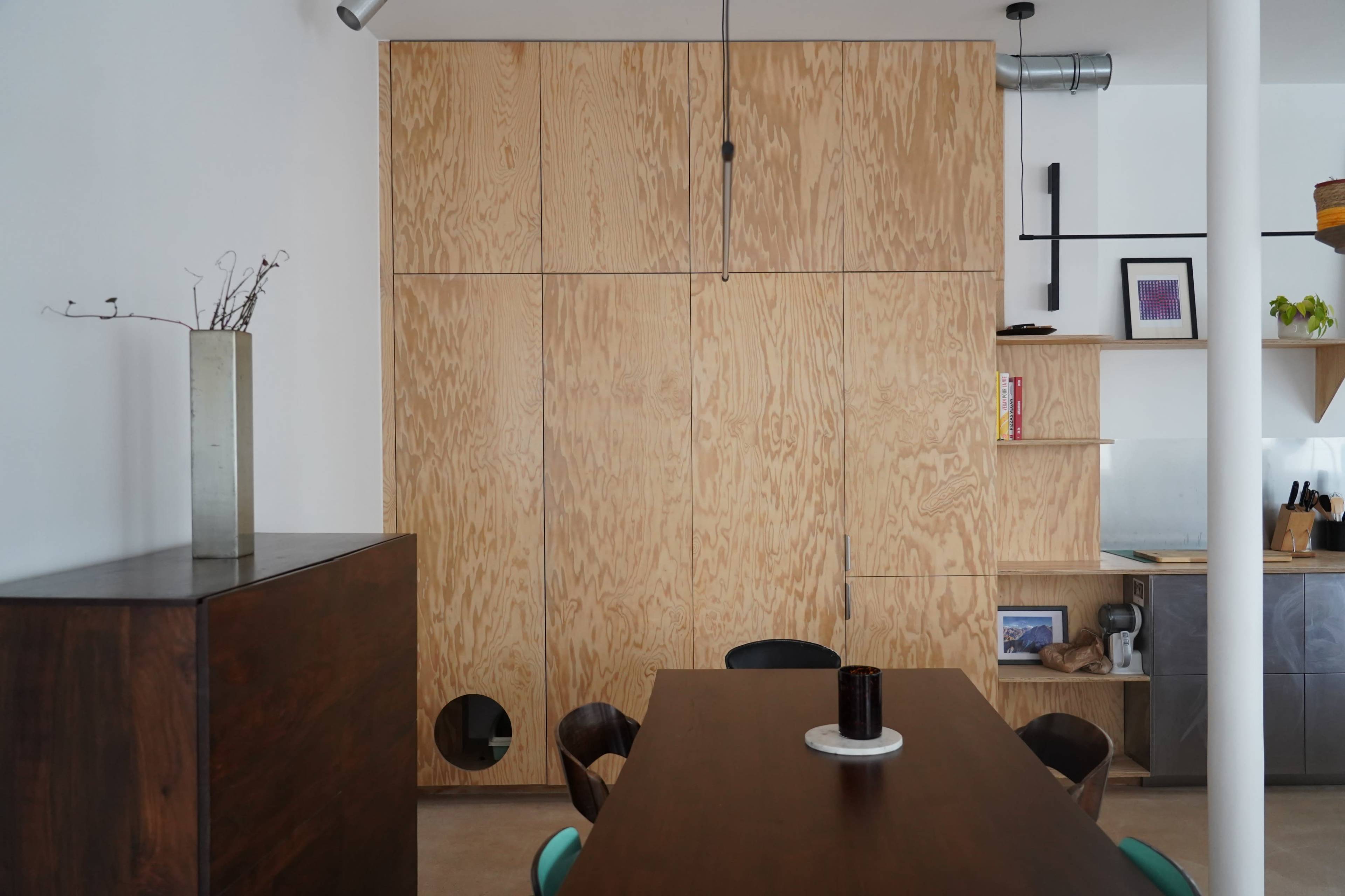 The image showcases a modern dining area with a large wooden table, surrounded by chairs, and a wall made of light wood panels featuring built-in shelves and storage.