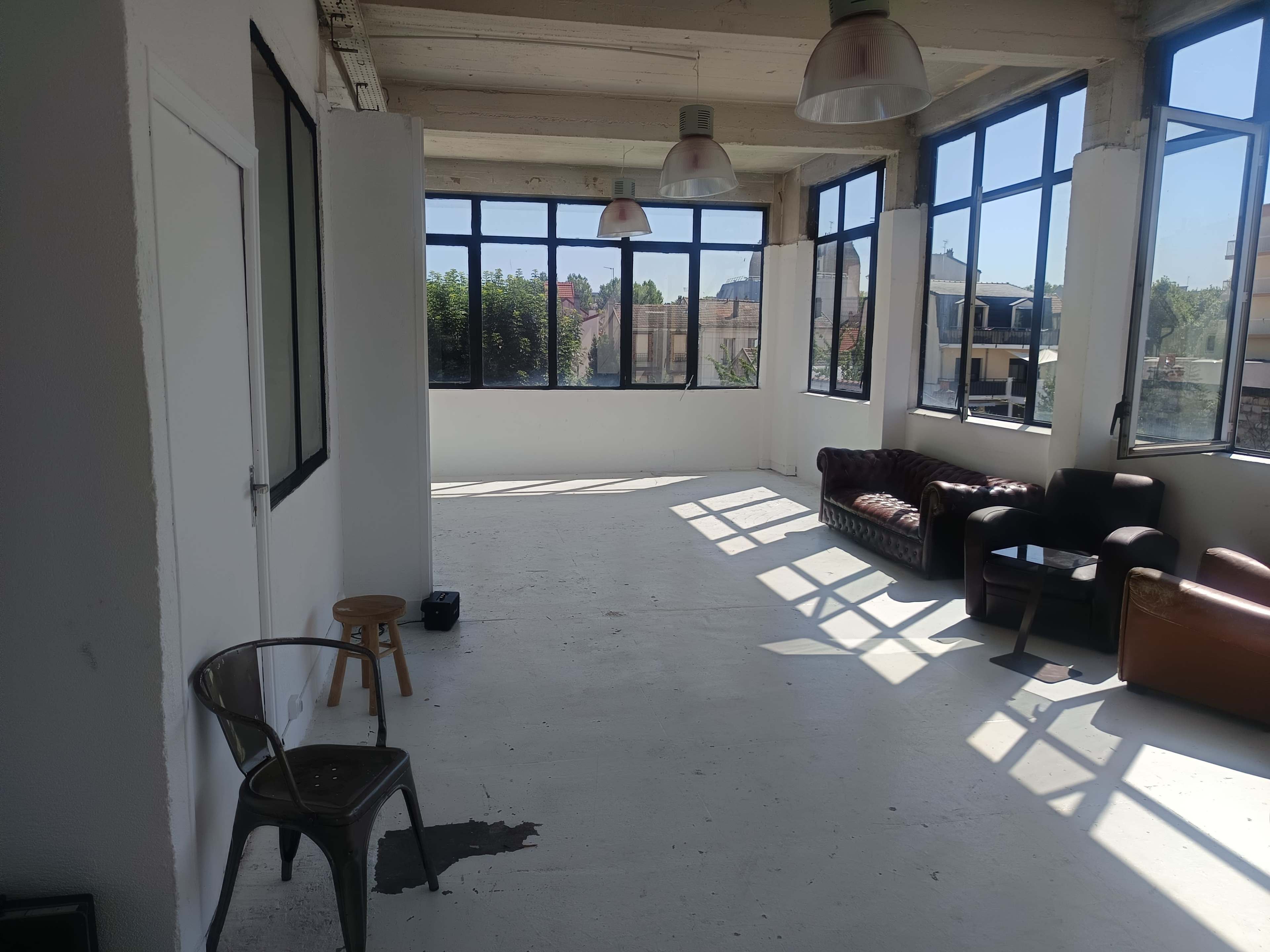 The image shows an interior space with large windows, showcasing bare white walls and a floor, featuring a brown leather sofa and a dark upholstered couch, along with a stool and a metal chair.