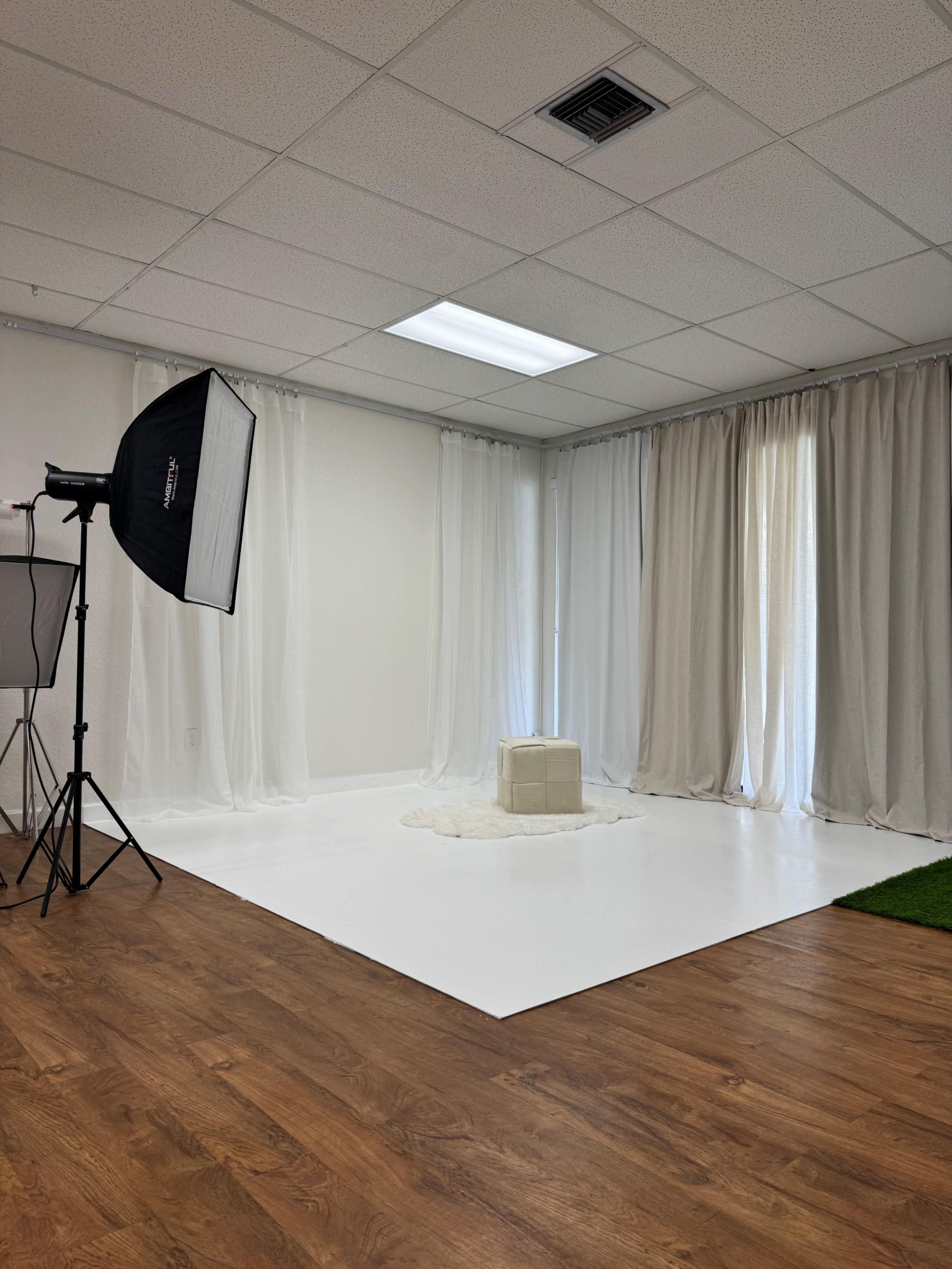The image shows a bright, minimalist photography studio with a white backdrop, a soft rug, and a light-colored box in the center.