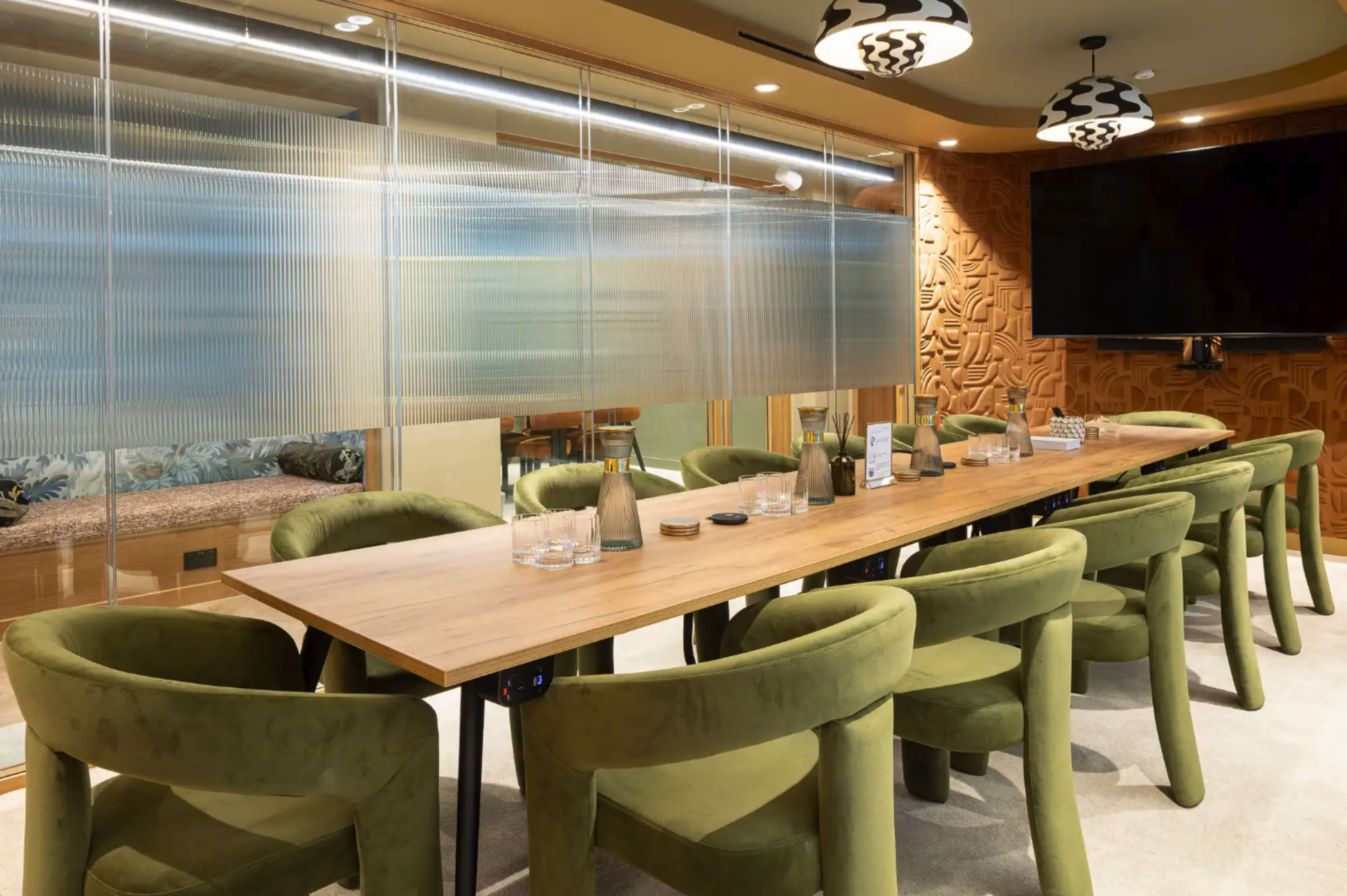 A long wooden table surrounded by green upholstered chairs is set in a modern meeting room with a large screen and decorative walls.