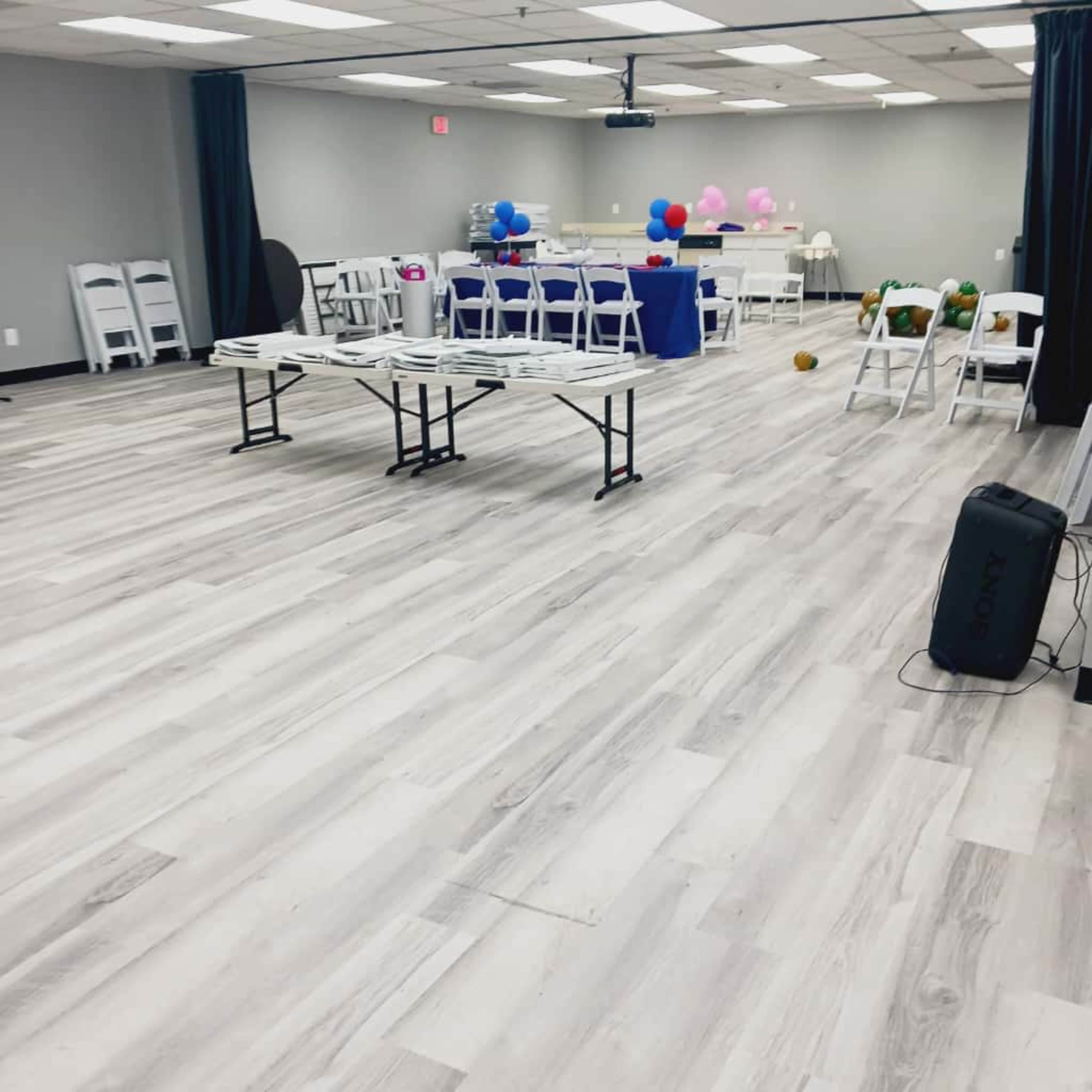 The image shows a spacious event room with light-colored flooring, foldable chairs arranged in rows, a table set up with decorations, and balloons in the background.