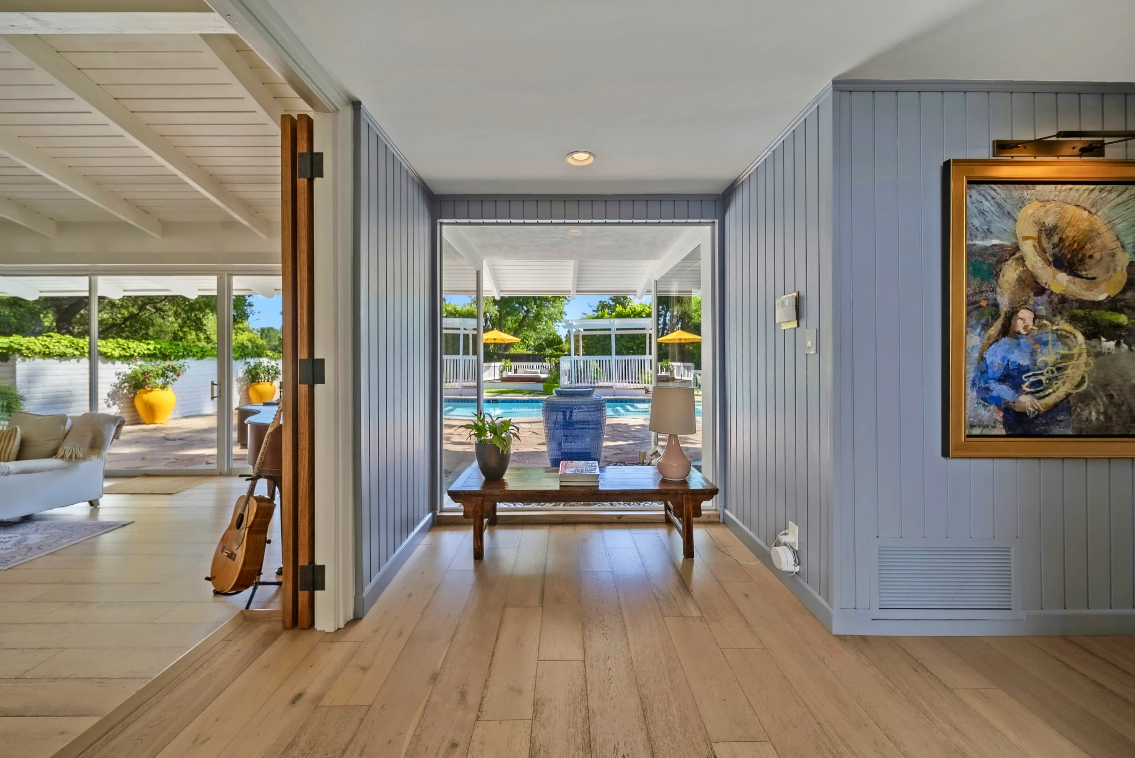 A hallway with light wood flooring leads to an outdoor area featuring a pool and patio furniture, framed by a blue paneled wall and a painting on the side.