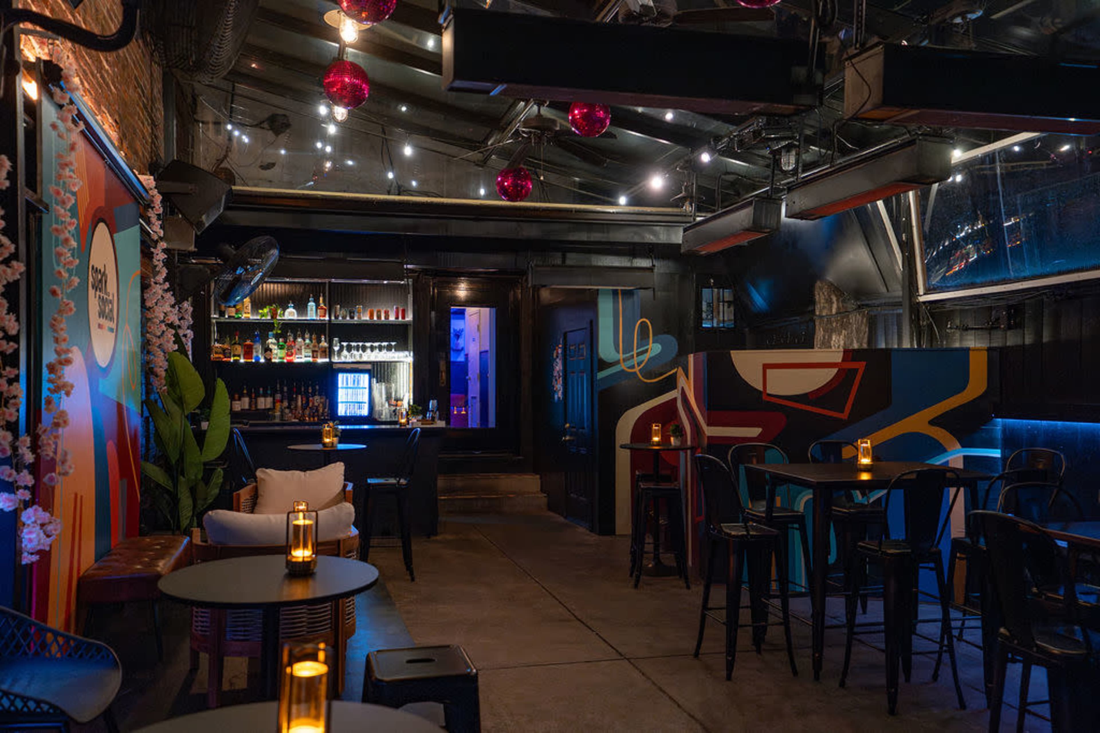 The image shows an interior view of a dimly lit bar featuring colorful murals, a bar area stocked with bottles, and several tables with candles.