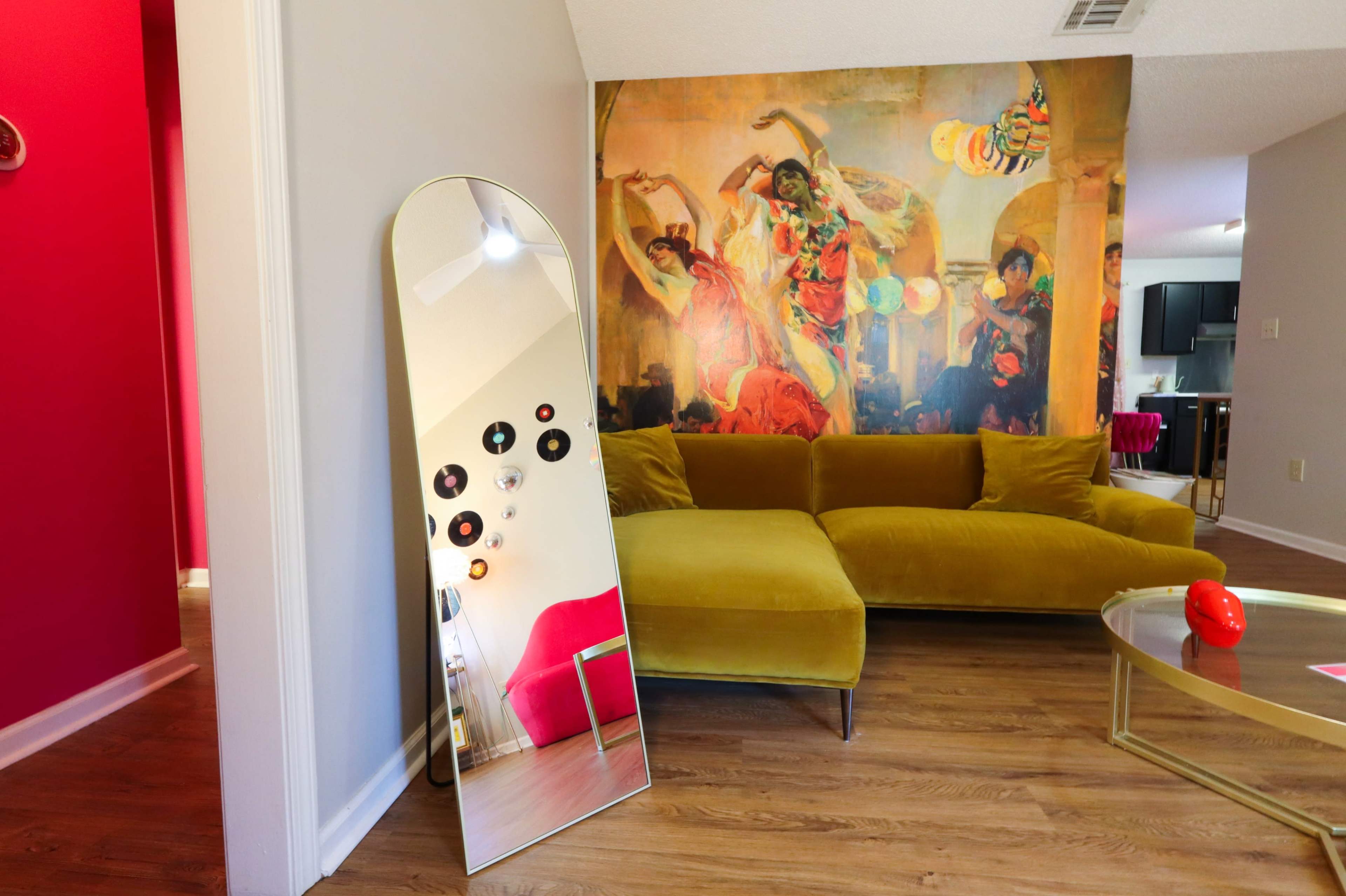 The image shows a living room with a large mirror reflecting a colorful wall mural of dancers, a mustard-colored sofa, and a pink chair.