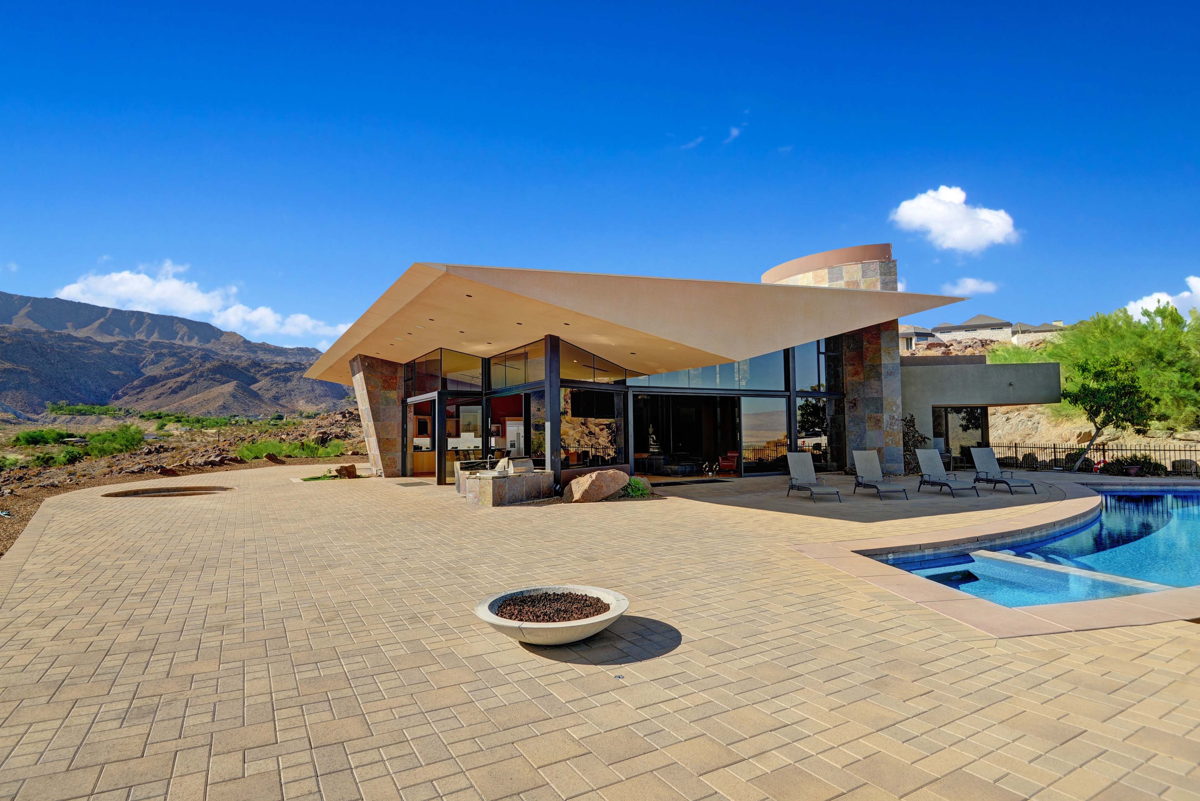 A modern, angular house is situated near a swimming pool, surrounded by rocky terrain and mountains under a clear blue sky.