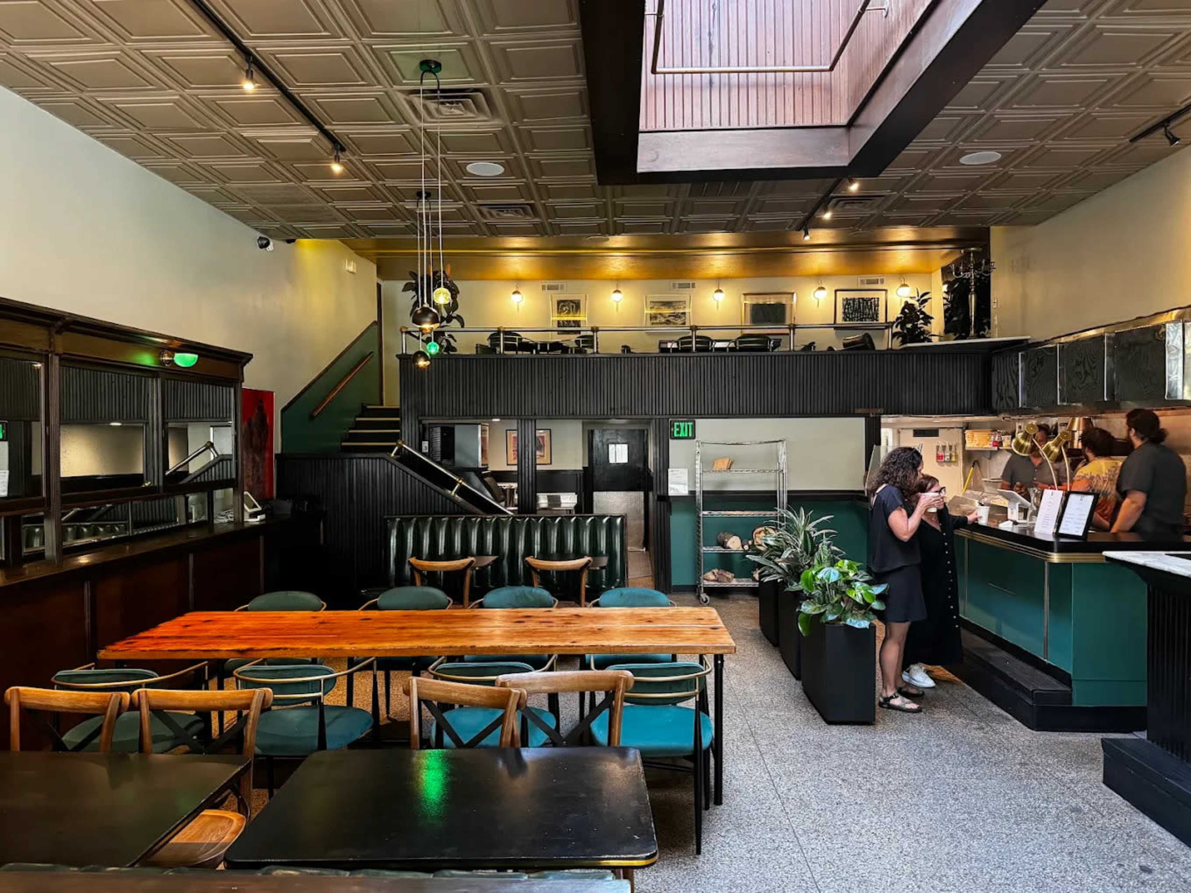 The image shows an interior view of a restaurant with a mix of seating arrangements, including dining tables and a bar area, under a skylight.