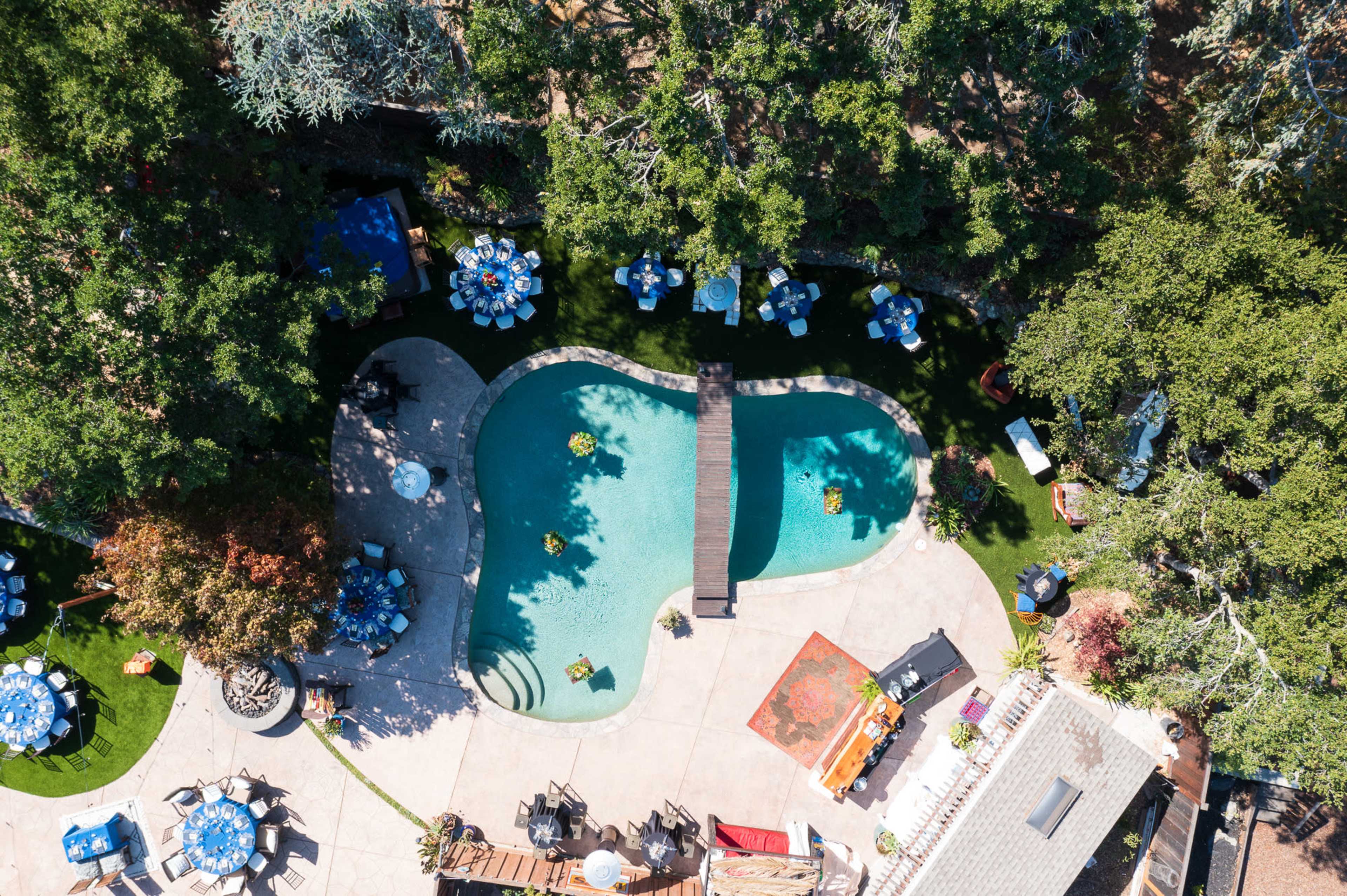 An aerial view of a landscaped backyard featuring a curvy swimming pool, a wooden bridge, and several tables arranged around it.