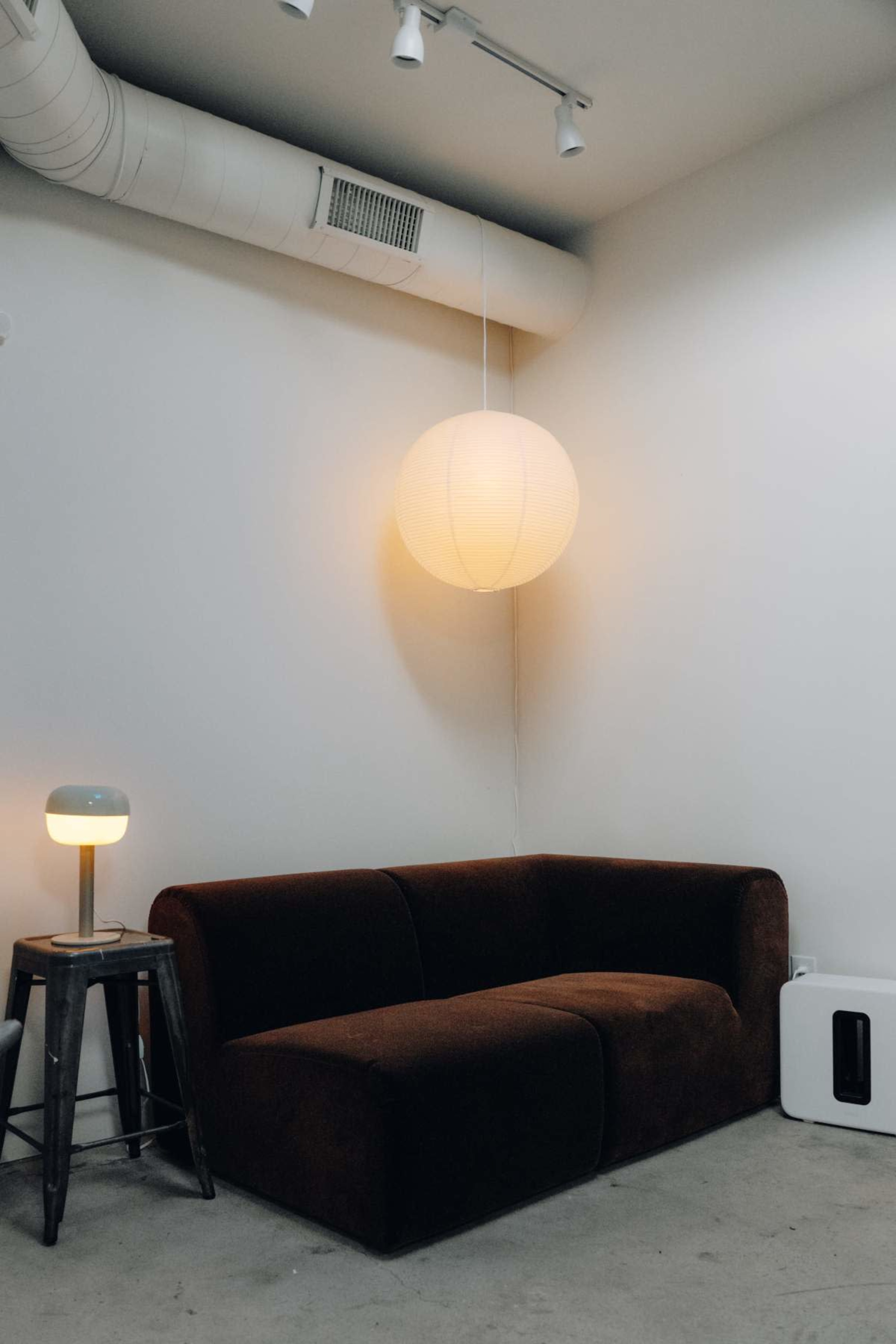 A brown sectional couch is positioned against a white wall, illuminated by a round hanging light above and a small table lamp nearby.
