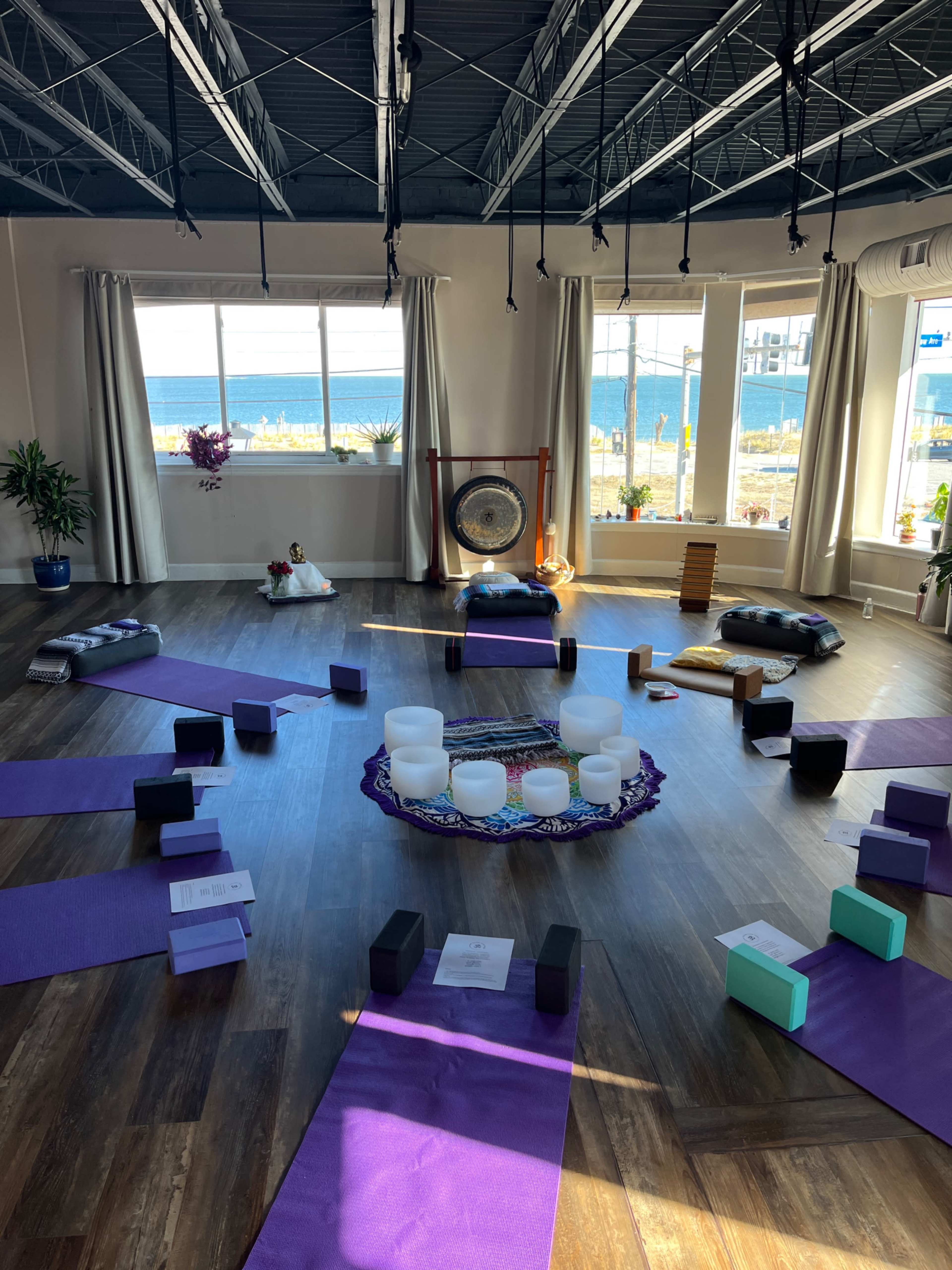 A spacious yoga studio with purple mats arranged in a circular pattern around a central altar, featuring a gong and several plants, with large windows showing a view of the ocean.