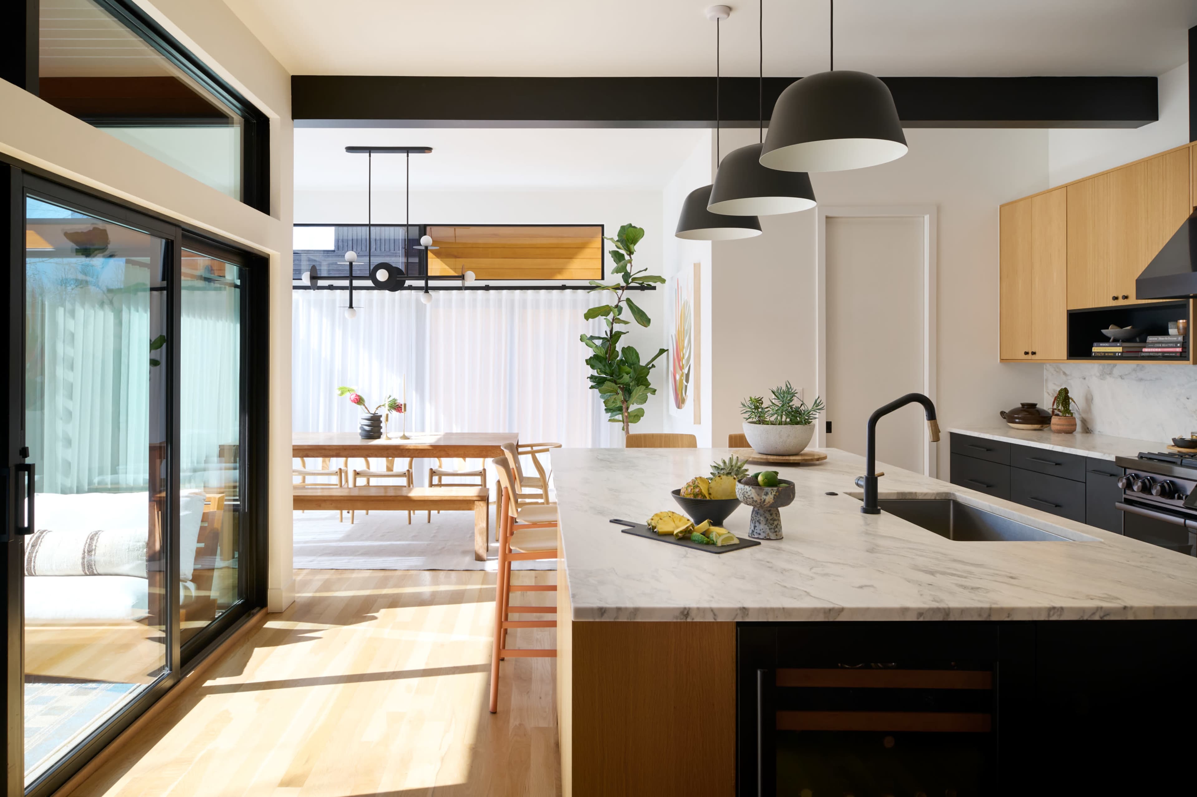 The image features a modern kitchen open to a dining area, with a marble island, wooden dining table, and large windows allowing natural light.