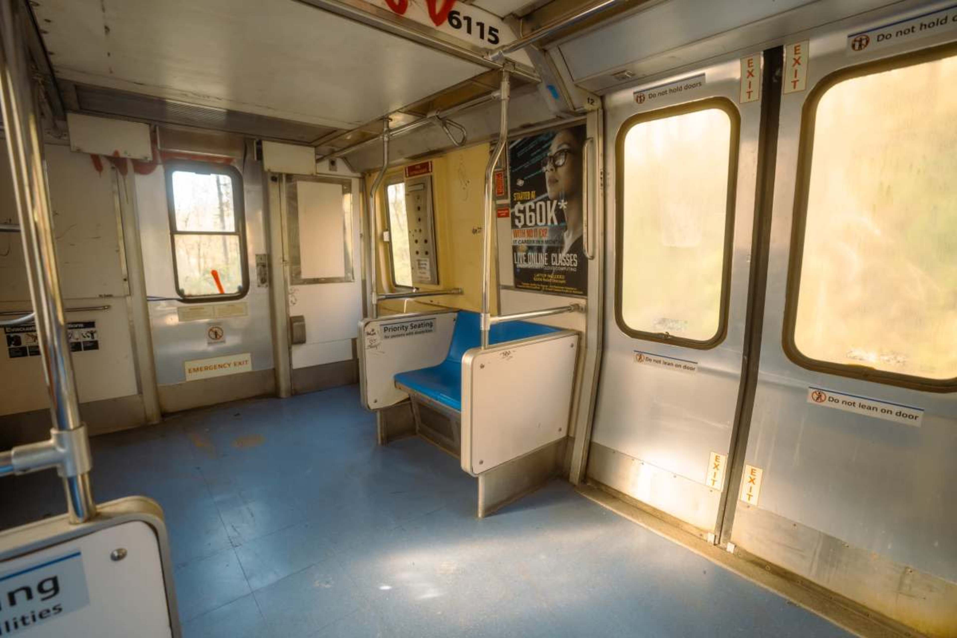 An empty subway car features a few seats along the walls and large windows allowing light to enter.