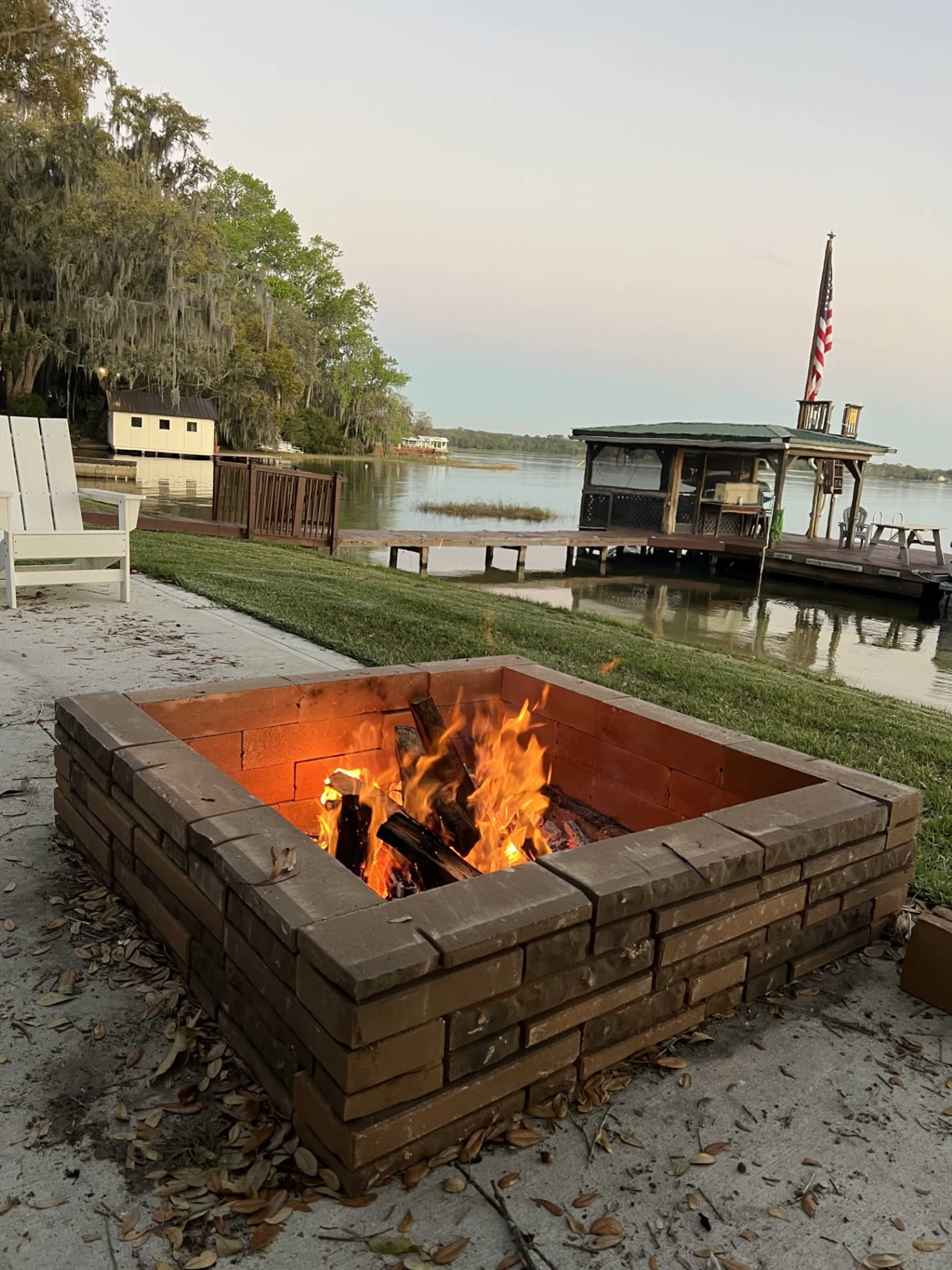 A brick fire pit with flames is located near a calm body of water, with a docked boat and a flag in the background.