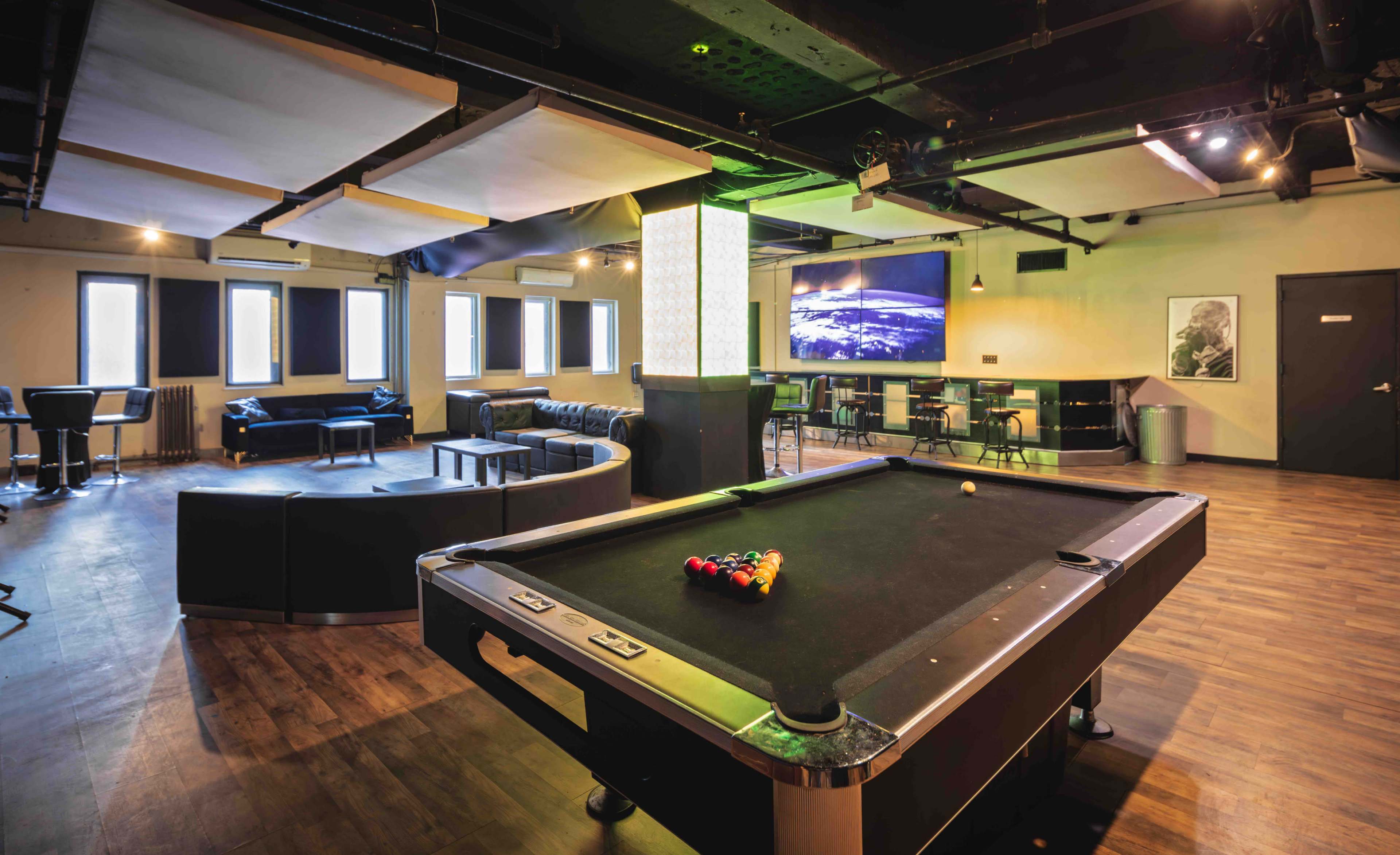 The image shows a modern lounge area featuring a pool table, comfortable seating, and a large screen displaying content.