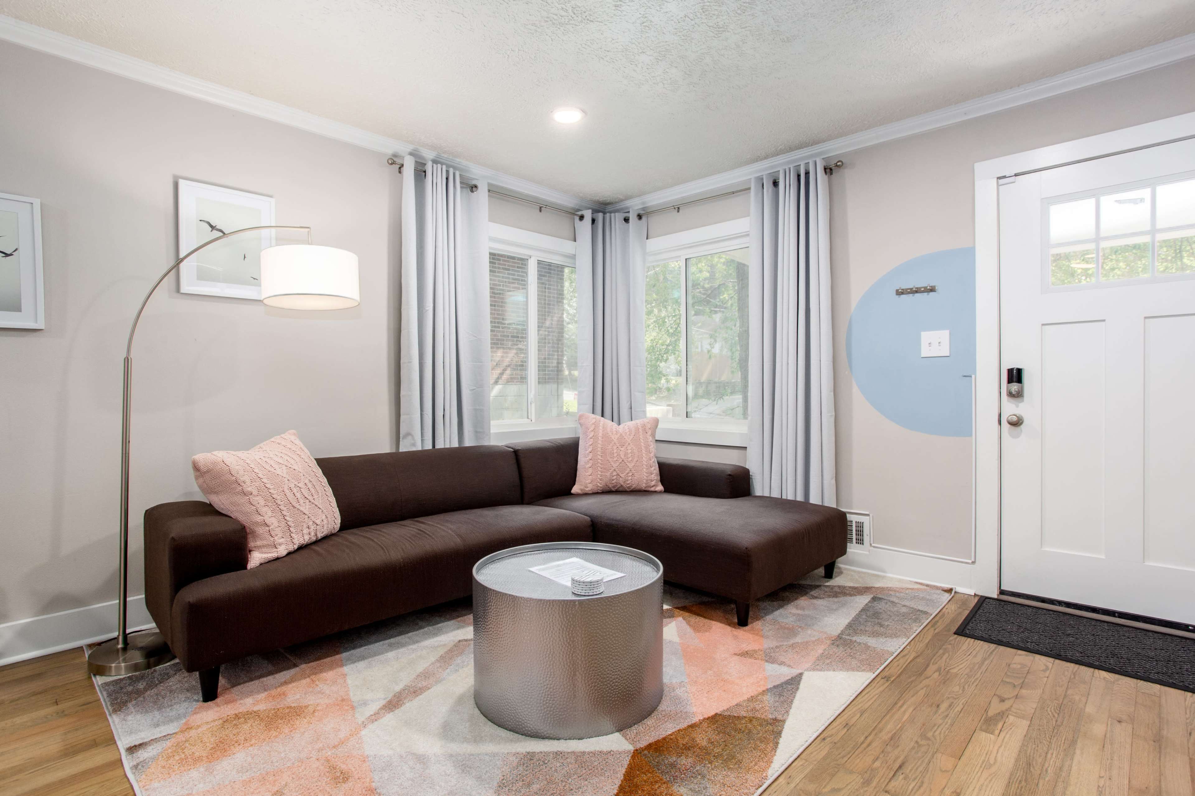 A modern living room featuring a brown sectional sofa, a circular glass coffee table, and large windows dressed with light gray curtains.