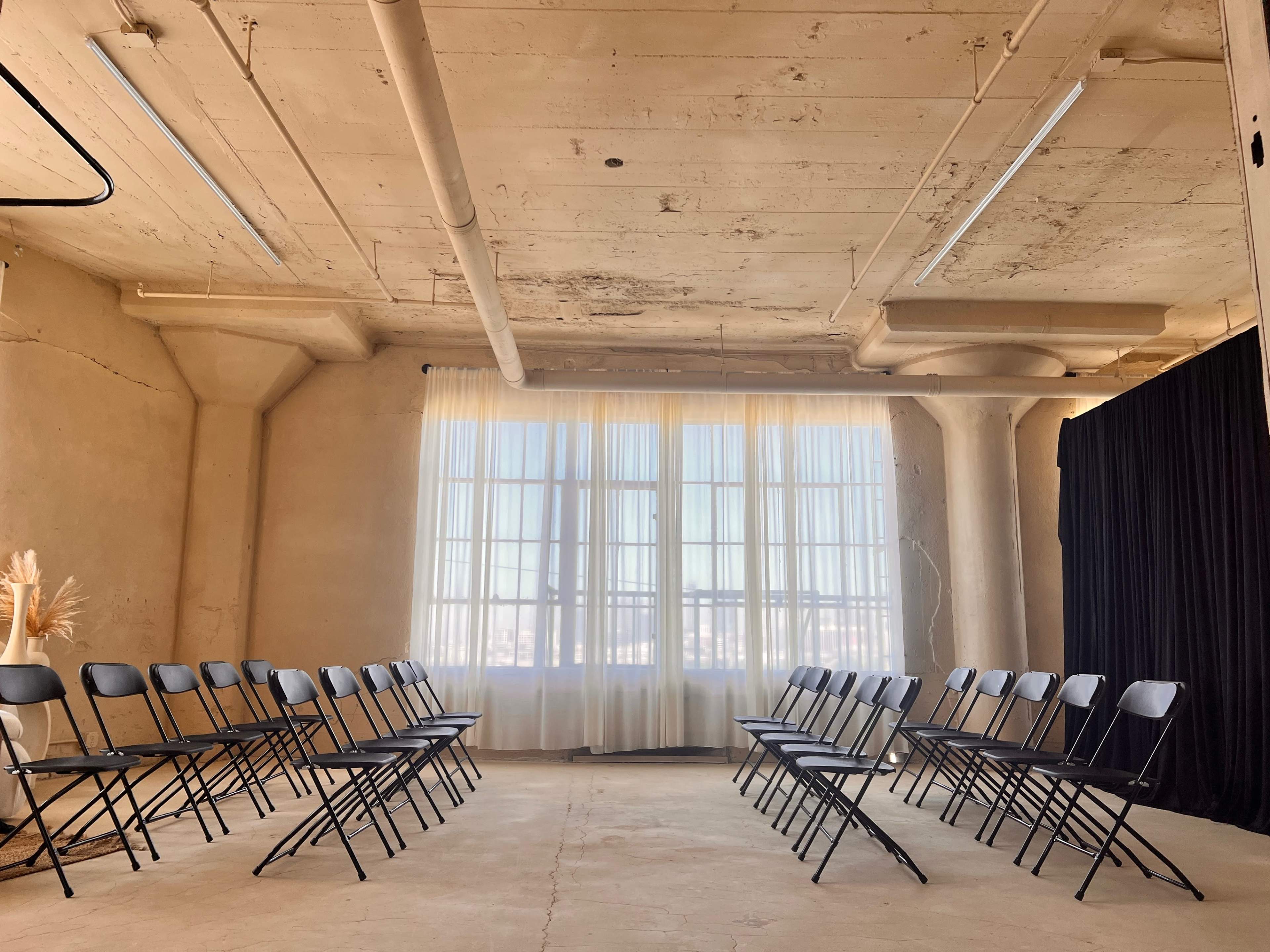 A room with a concrete floor features two rows of folding chairs facing each other, near large windows with sheer curtains and a black backdrop.
