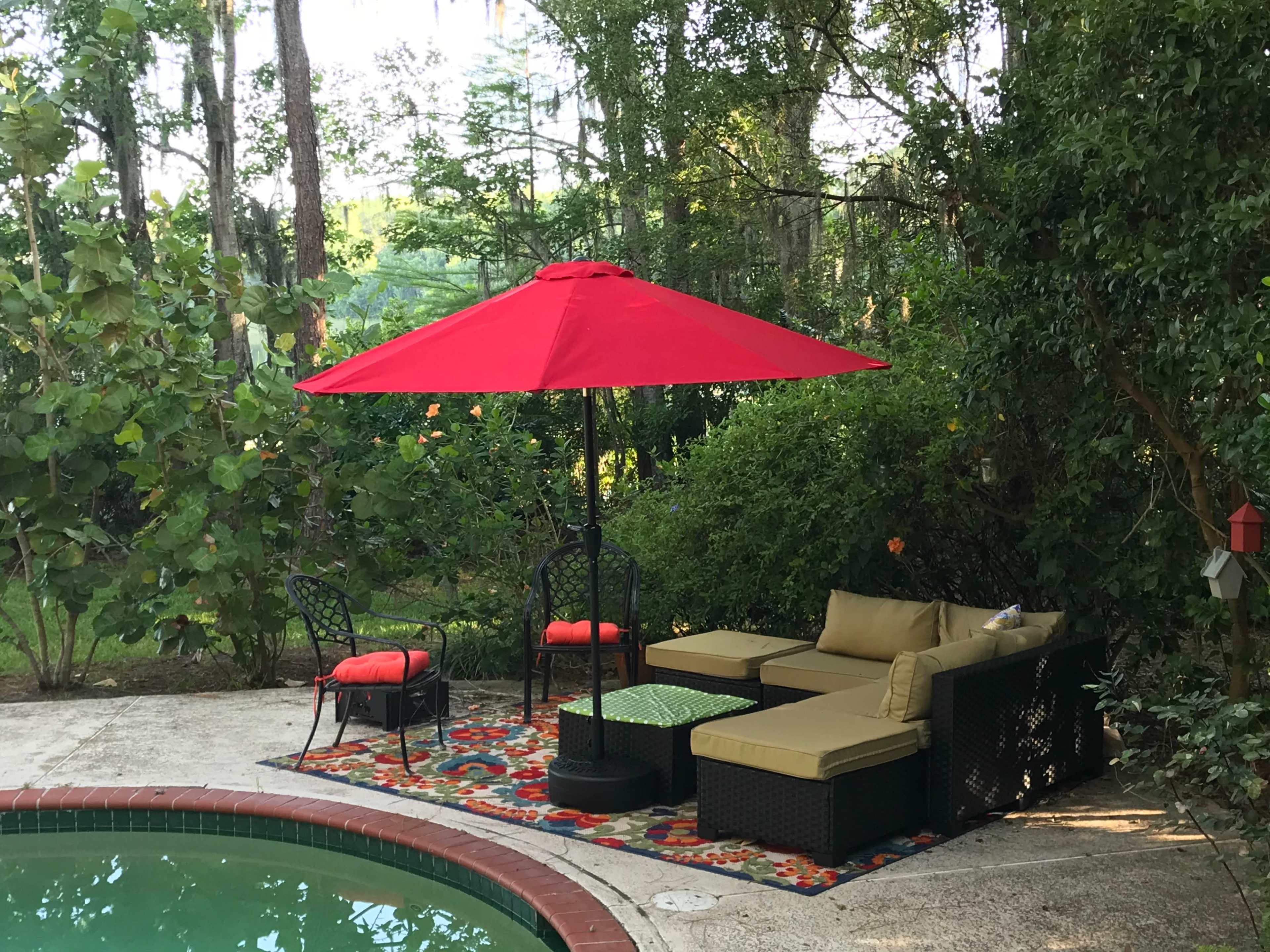A red umbrella shades a seating area with a sofa and chairs beside a pool surrounded by greenery.