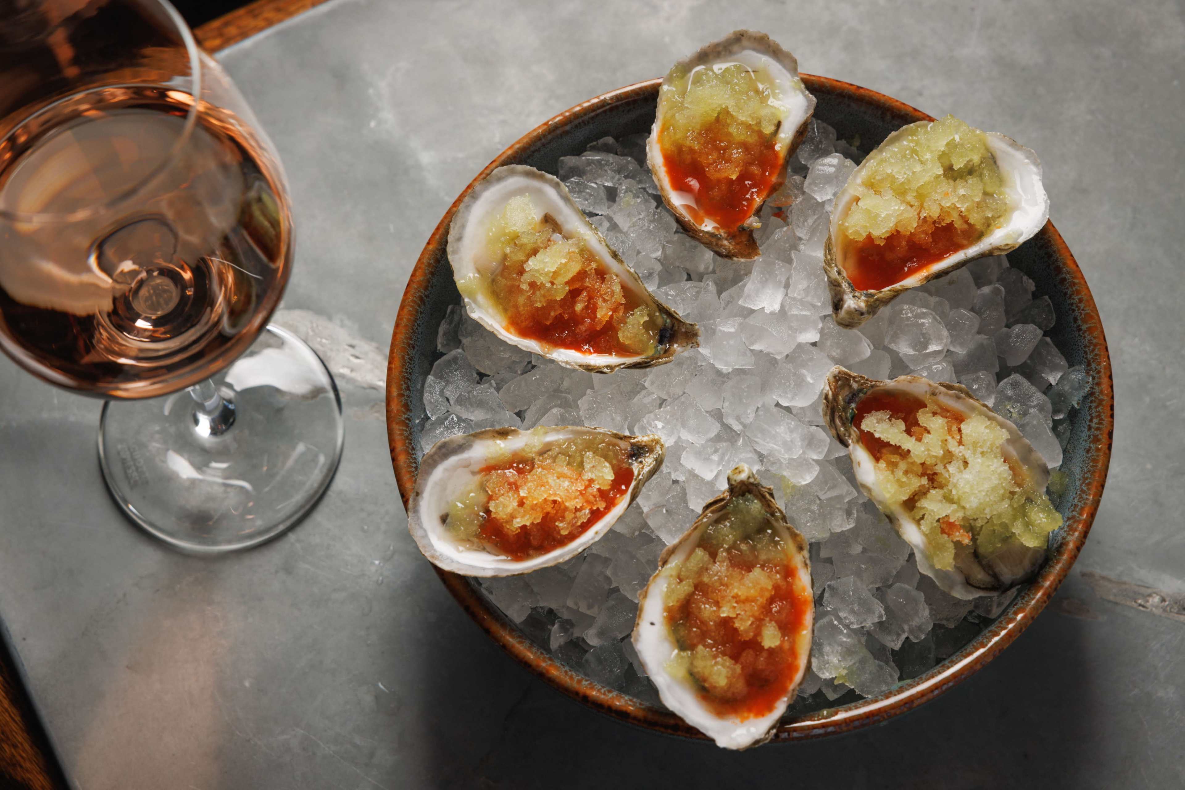 A bowl of six oysters topped with a garnished mixture sits on ice beside a glass of rosé wine.