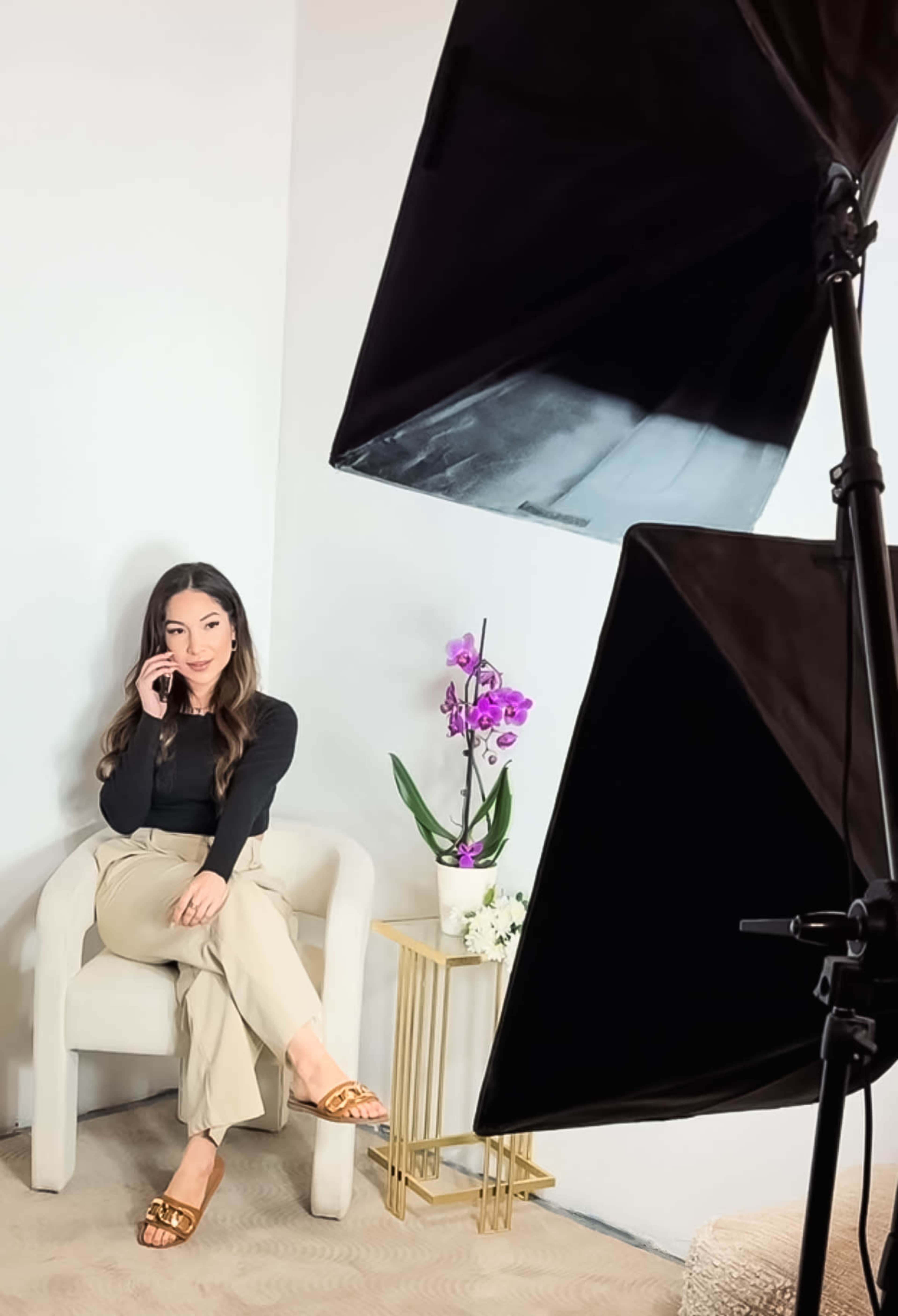 A woman sits on a chair in front of a light setup, holding a phone, with an orchid arrangement beside her.