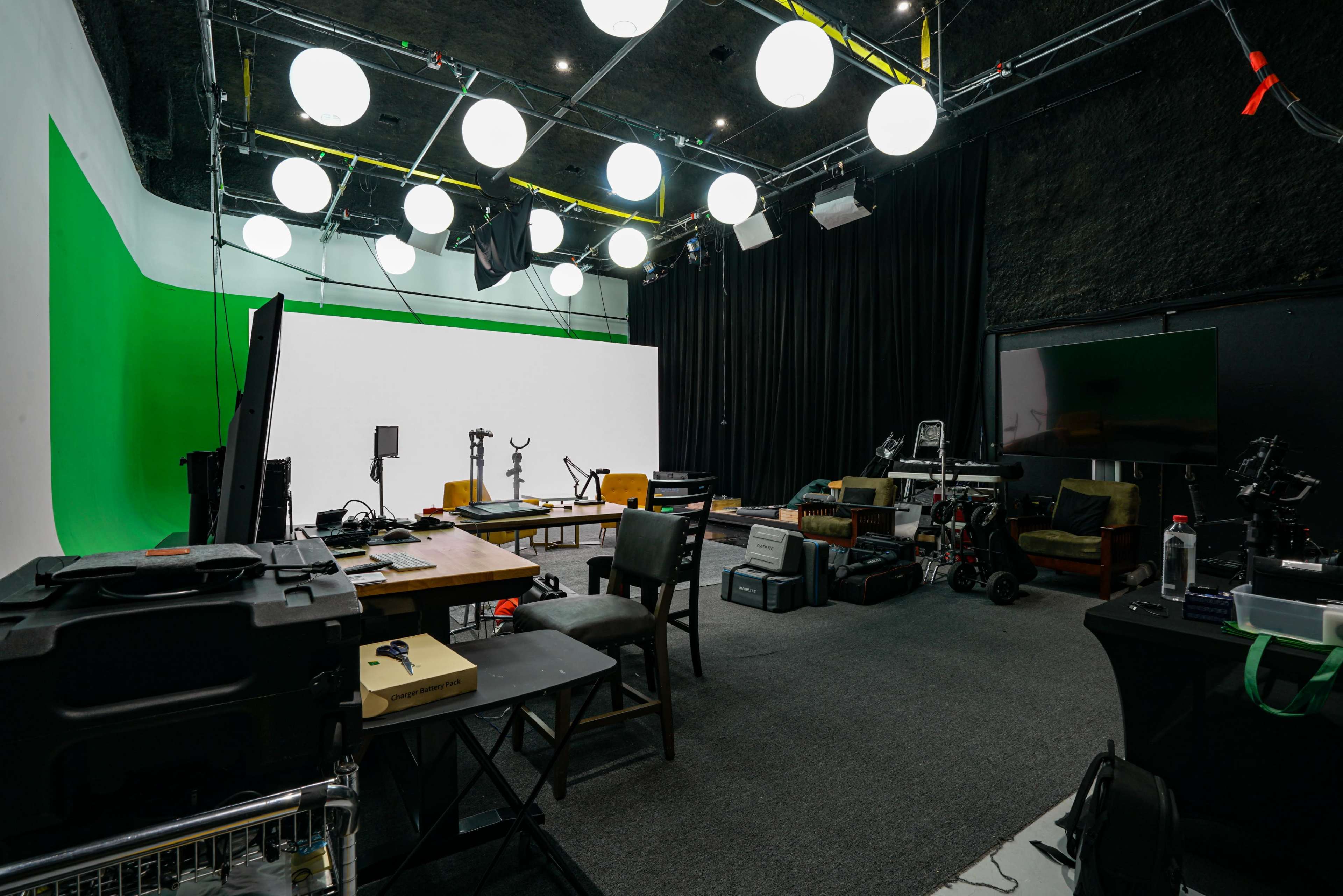 The image shows a production studio workspace with a green screen, various equipment, and tables arranged around a spacious area.