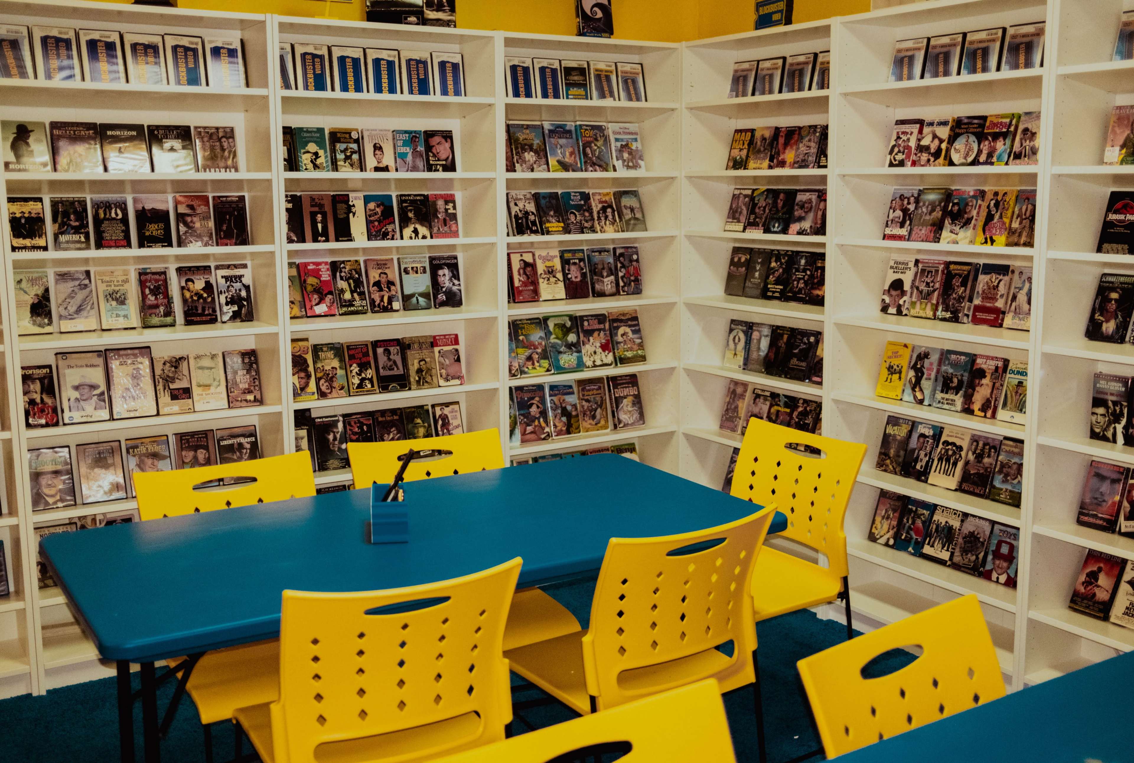 A brightly colored room features a collection of VHS tapes neatly organized on white shelves, alongside blue tables and yellow chairs arranged for seating.