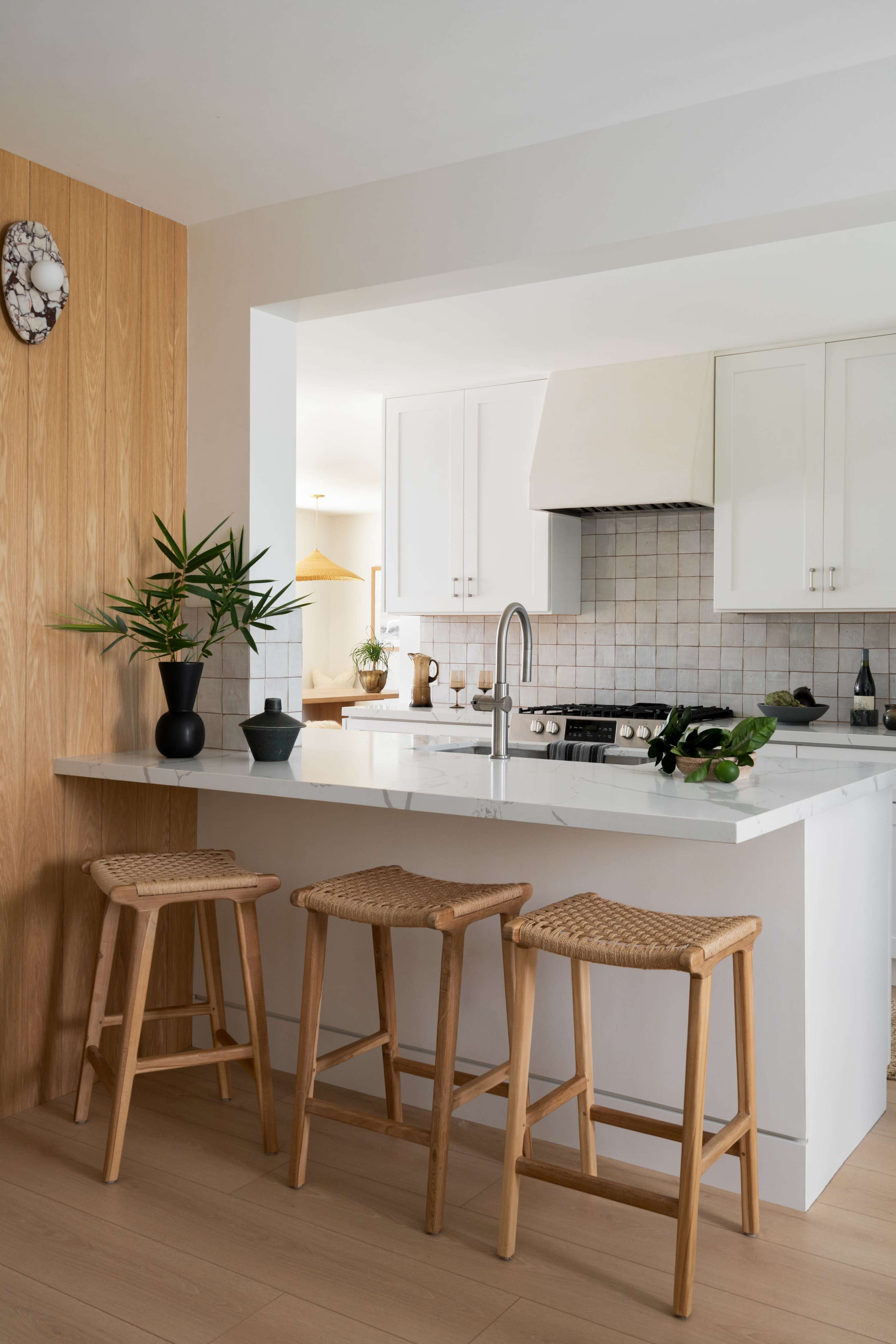 The image shows a modern kitchen with a bar area featuring three wooden stools, potted plants, and white cabinetry.