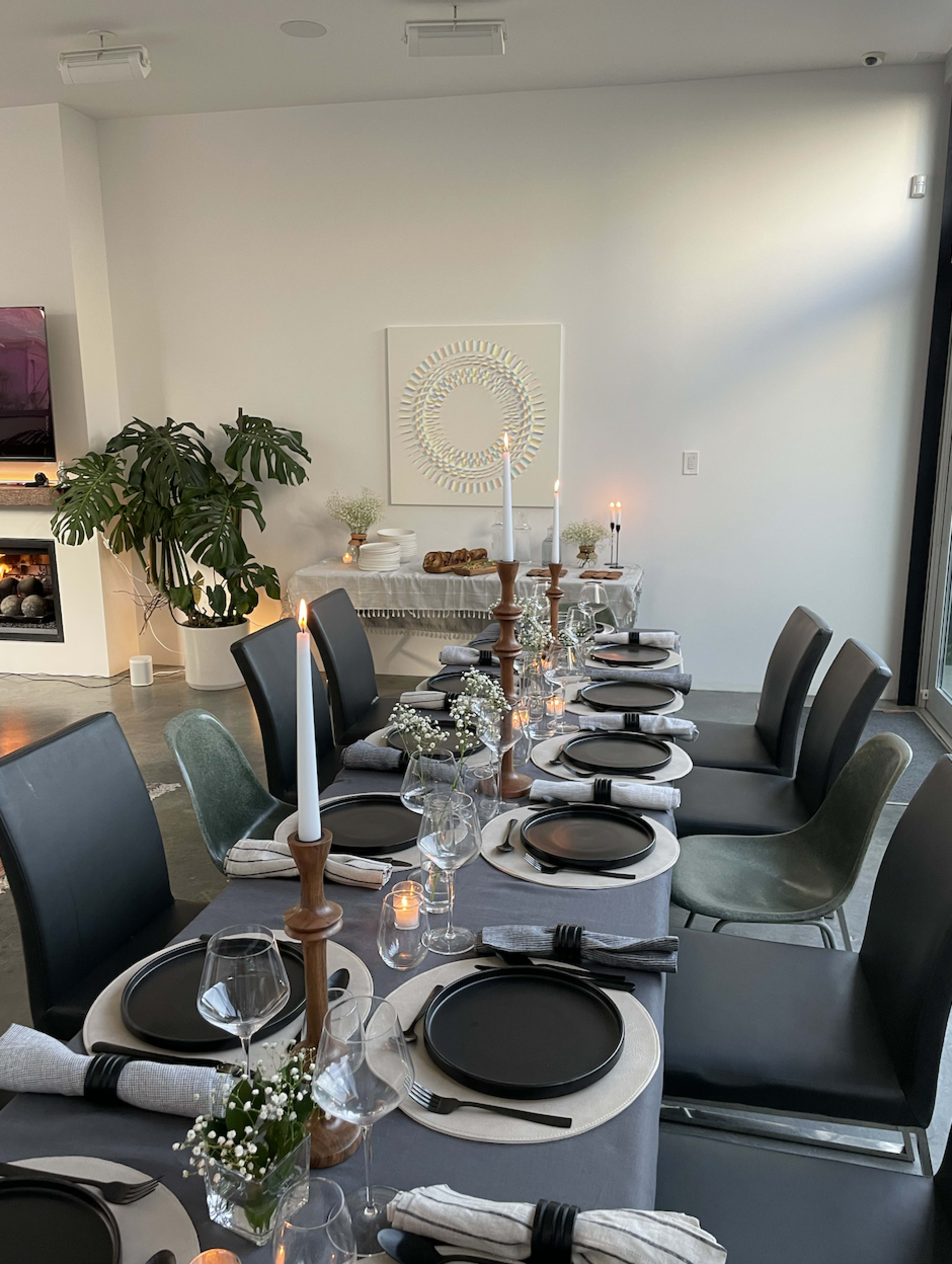A long dining table is set with black plates, glassware, and candles, surrounded by modern chairs in a bright room with a decorative wall piece and plant in the background.