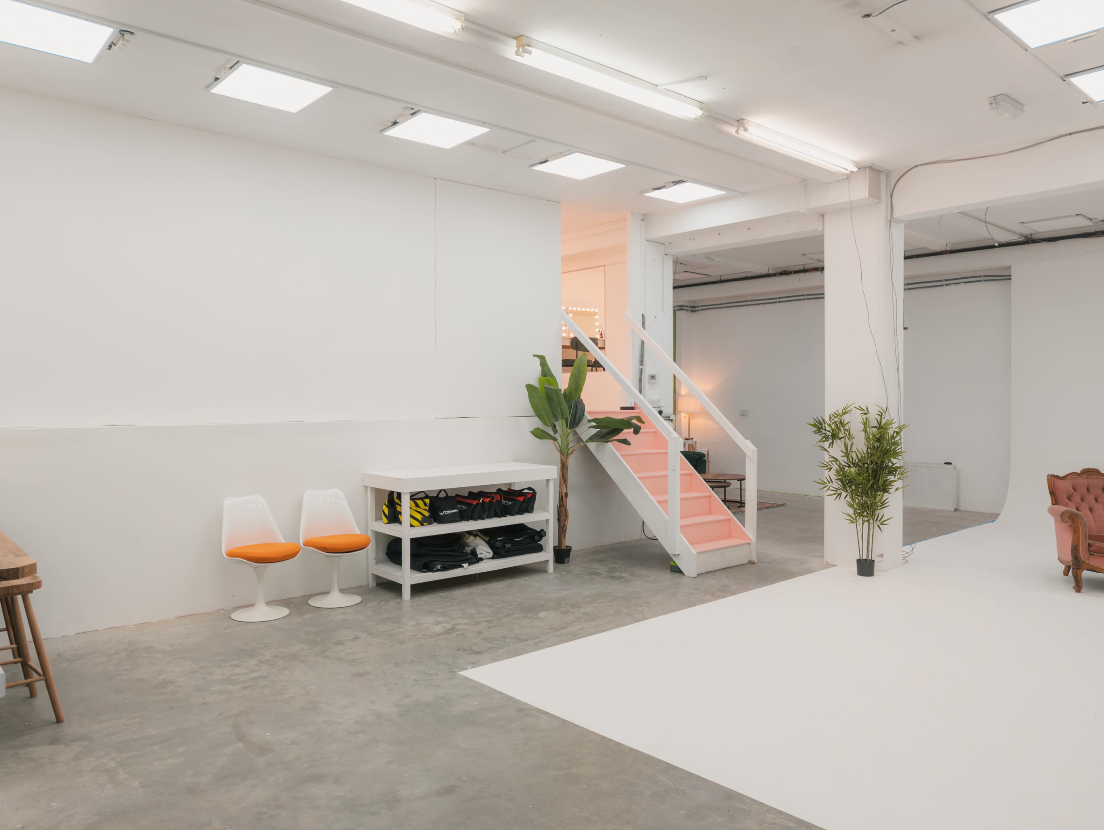 A bright, minimalist studio space with a staircase leading to a higher level, featuring a few chairs, plants, and a shelf with black and yellow items.