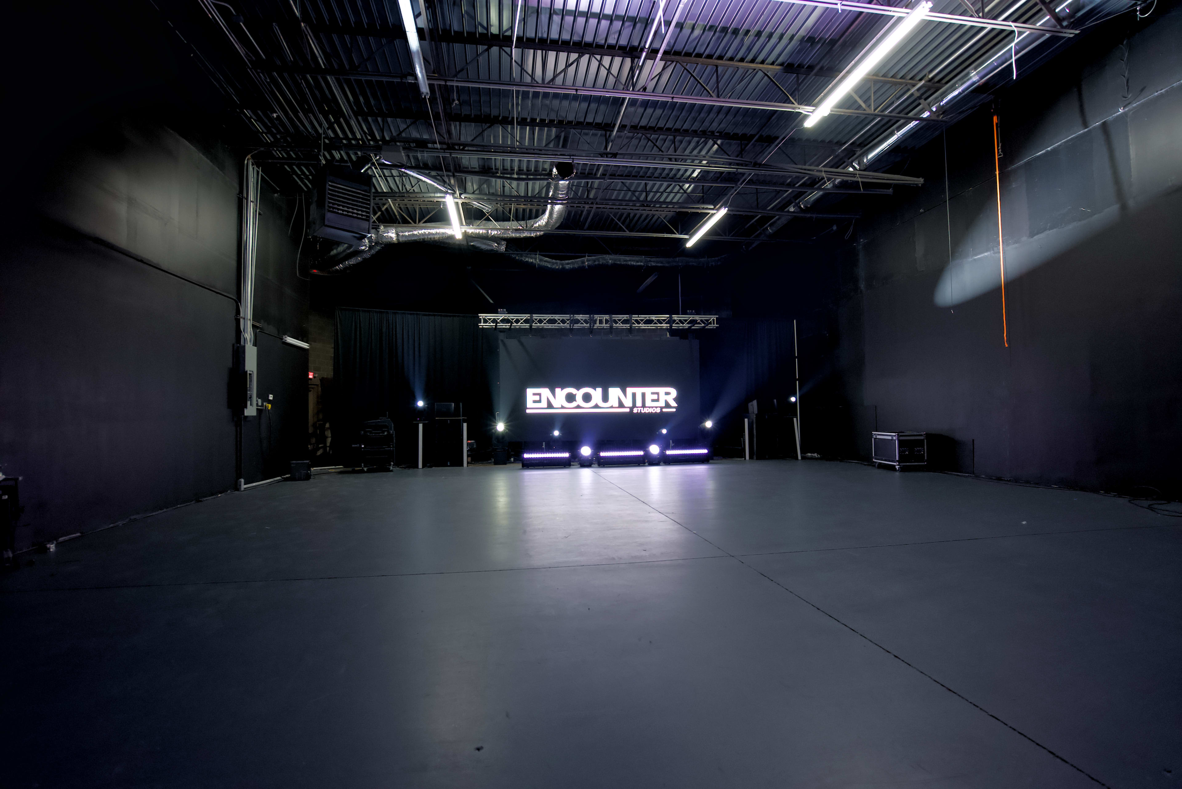 The image shows a large, empty warehouse-like space with a dark background and bright lights illuminating a stage featuring the word "ENCOUNTER."