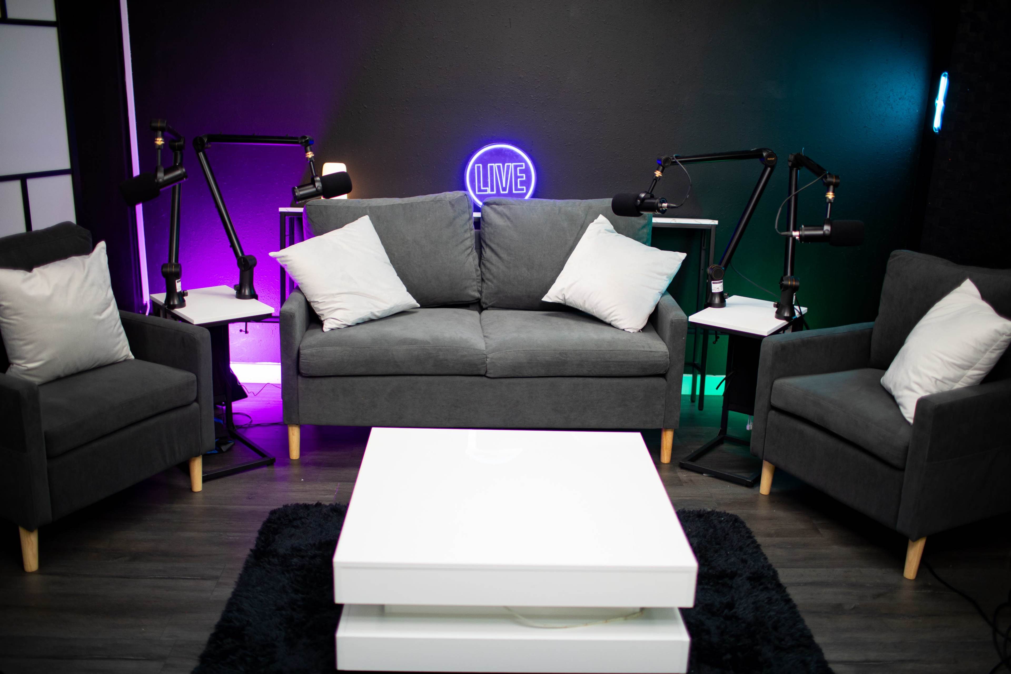 The image shows a modern, minimalist seating area with a gray couch, two matching armchairs, and a white coffee table, all set against a dark wall featuring a neon "LIVE" sign and microphone stands.