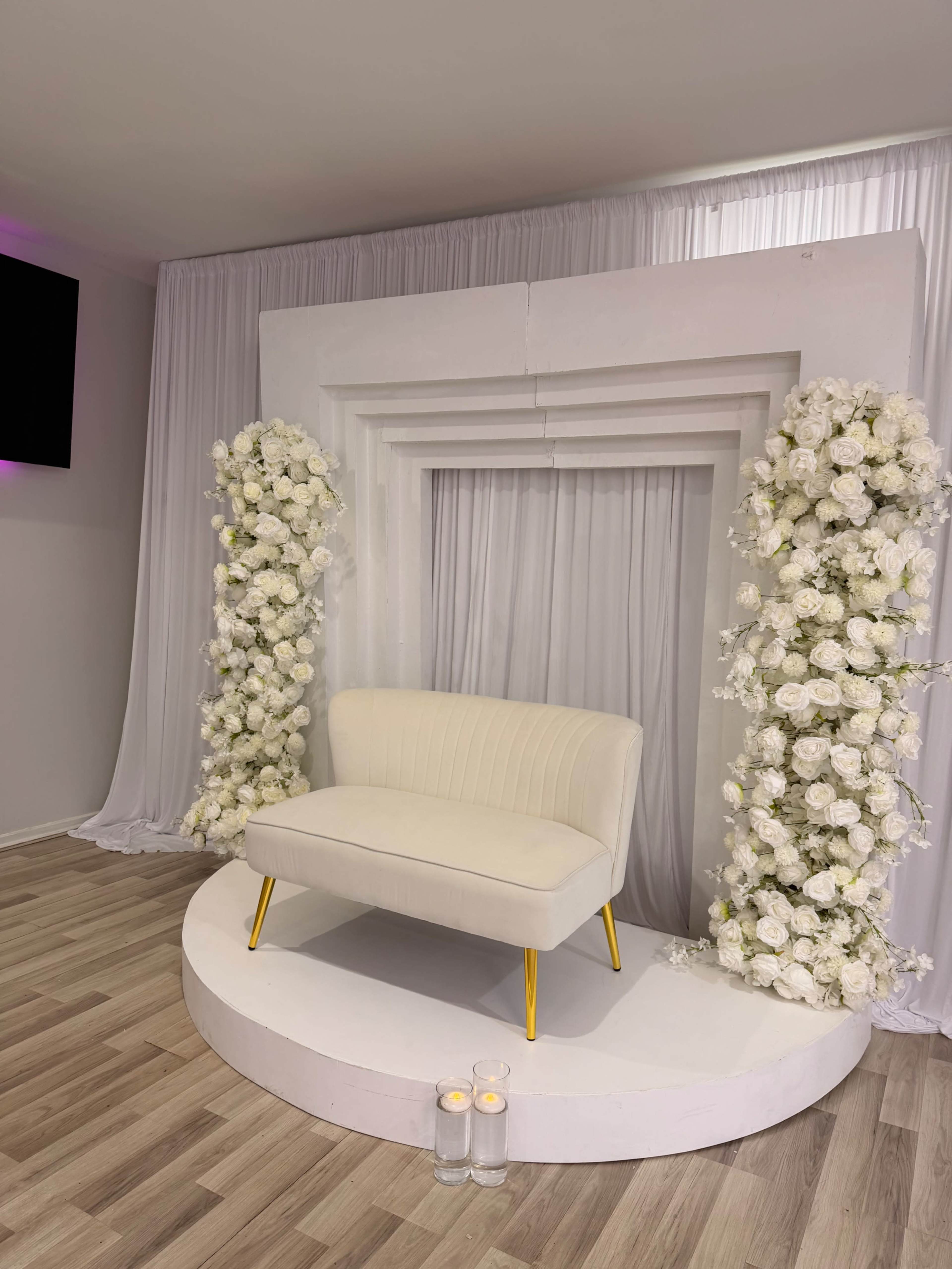 The image shows a small white seating area with a round pedestal surrounded by floral arrangements and draped curtains in a softly lit room.