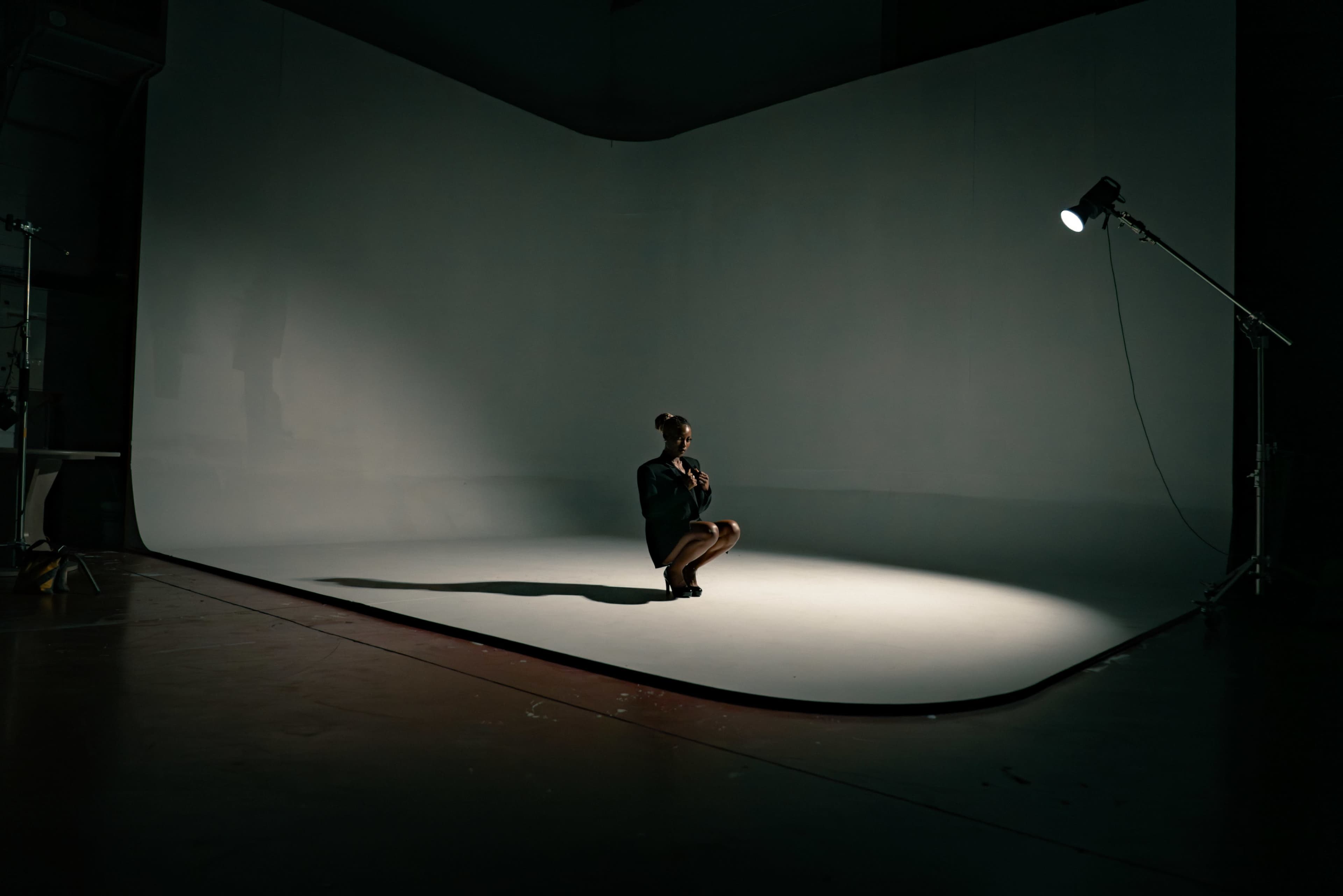 A solitary figure sits on a studio floor under a spotlight, surrounded by a blank backdrop.