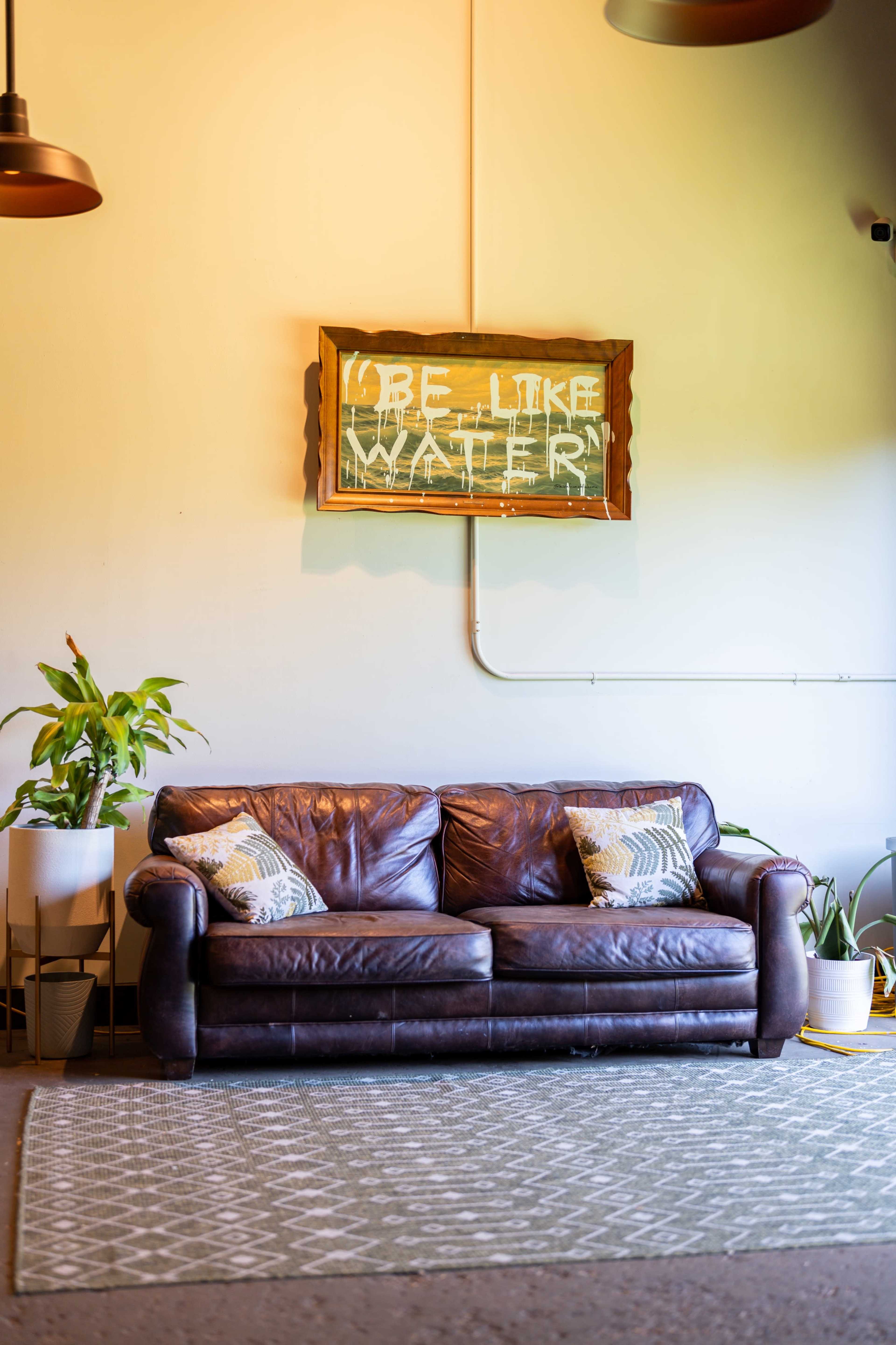 A brown leather sofa with decorative pillows sits on a patterned rug beneath a framed sign that reads "BE LIKE WATER," accompanied by potted plants.