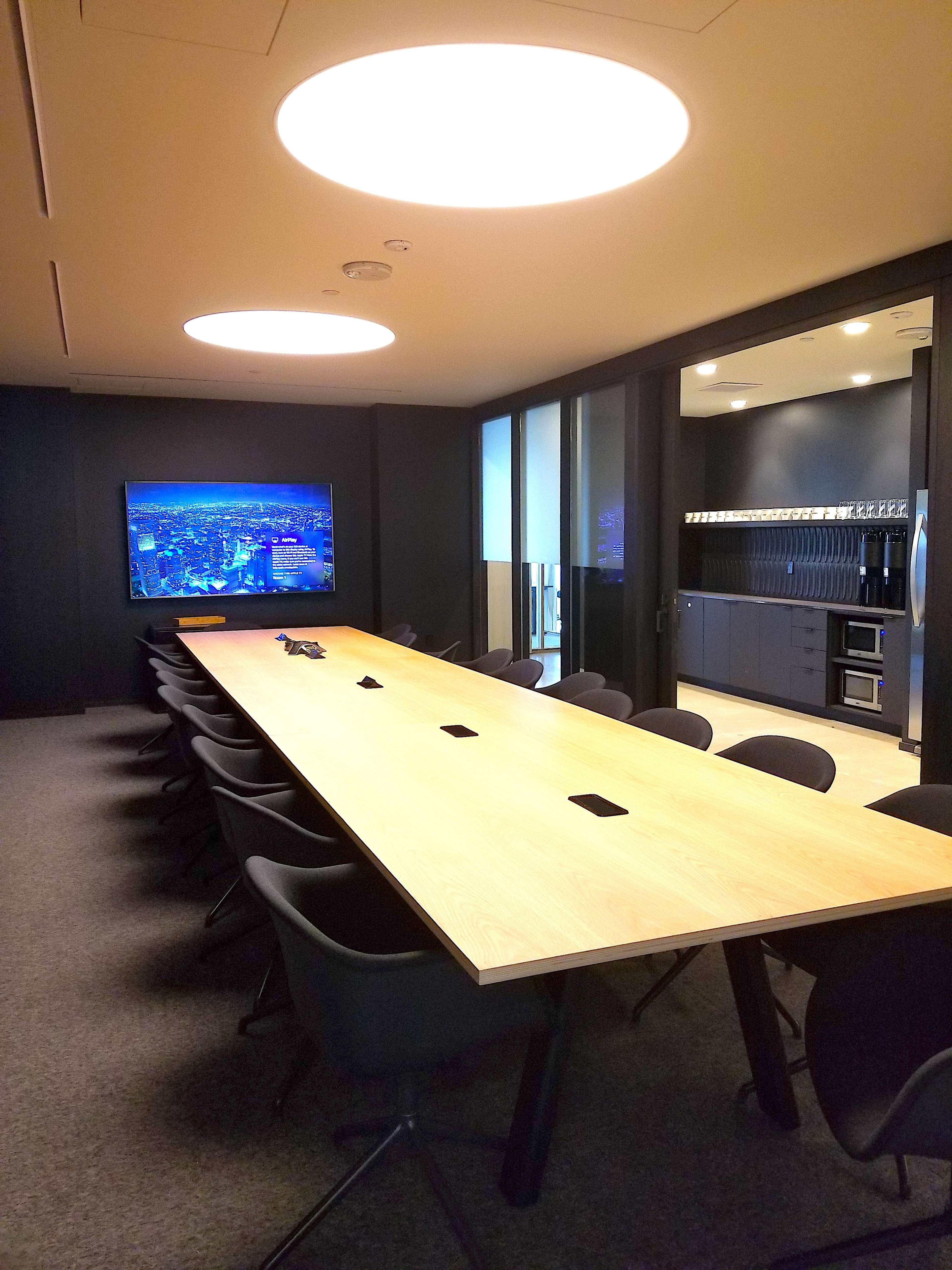 A modern conference room with a long wooden table, black chairs, and a large screen displaying graphics, along with a kitchenette in the background.