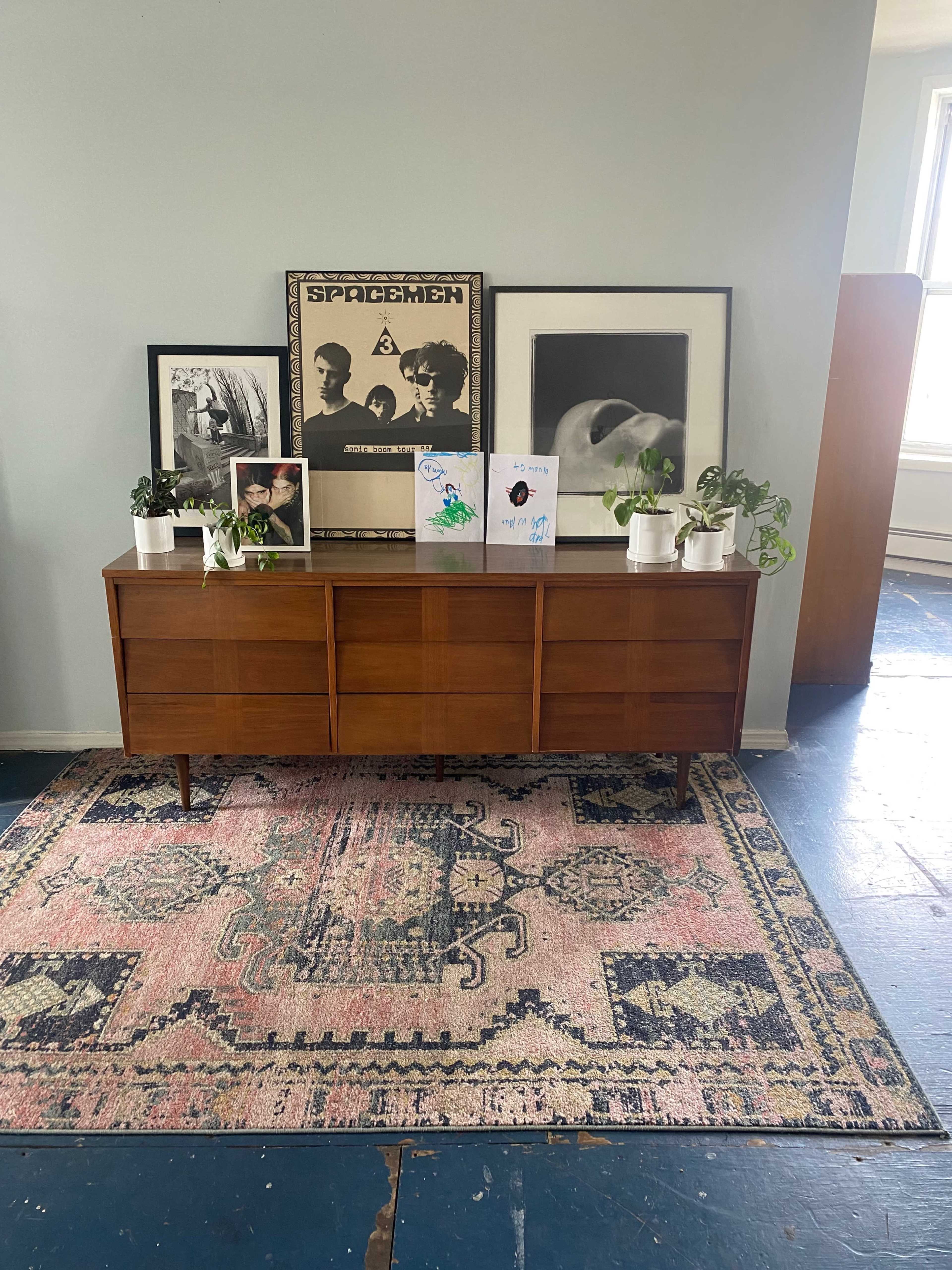 A wooden dresser sits against a light blue wall, adorned with framed artwork and small potted plants on top, with a patterned rug below.