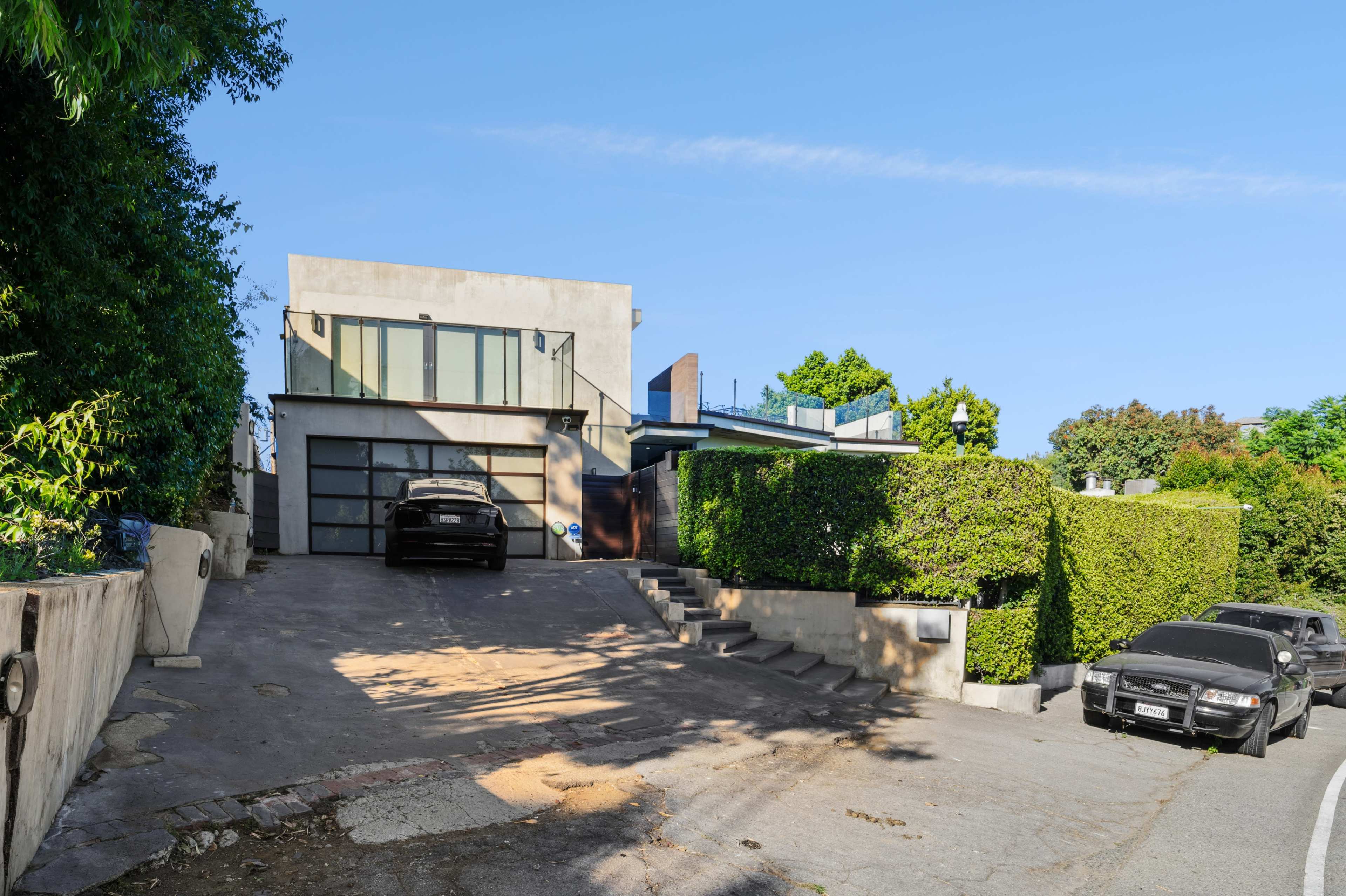 A modern house set on a sloped driveway, flanked by greenery, with two cars parked in front of a garage.