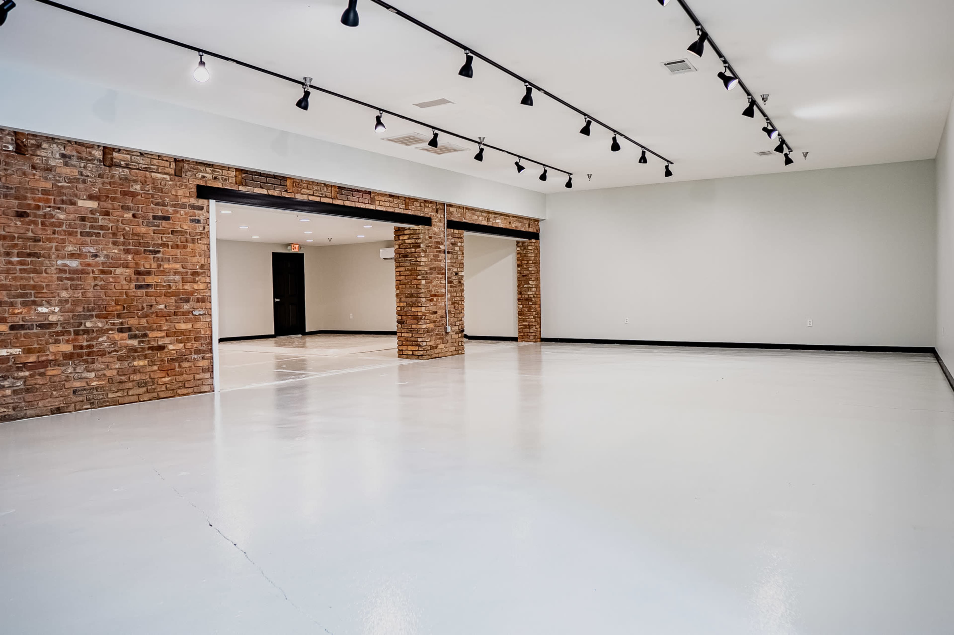 A spacious, empty room with white floors, exposed brick columns, and track lighting on the ceiling.