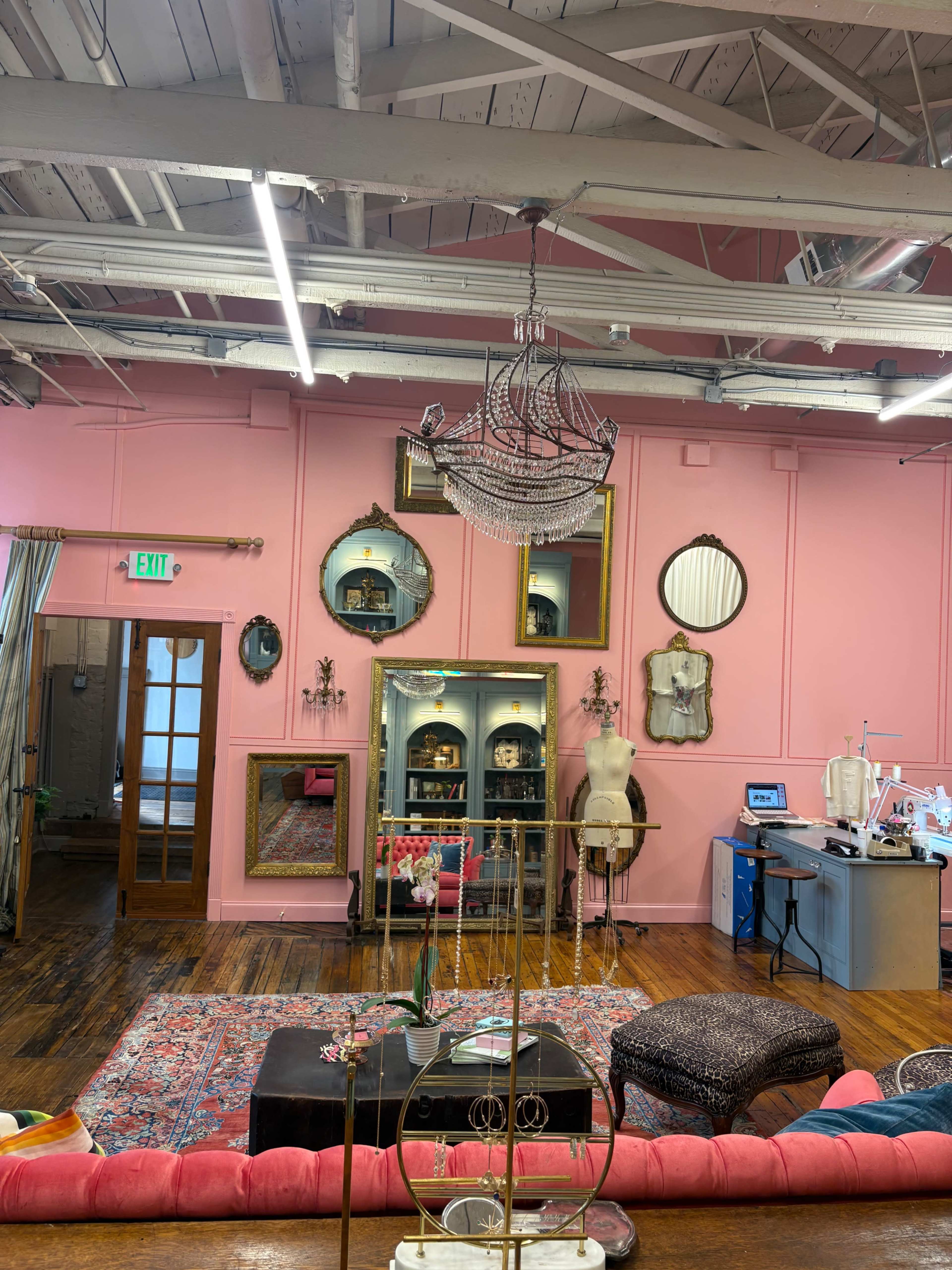 The image shows a brightly colored pink-walled room featuring a chandelier, decorative mirrors, a vintage mannequin, a large rug, and various furniture pieces arranged around a central seating area.