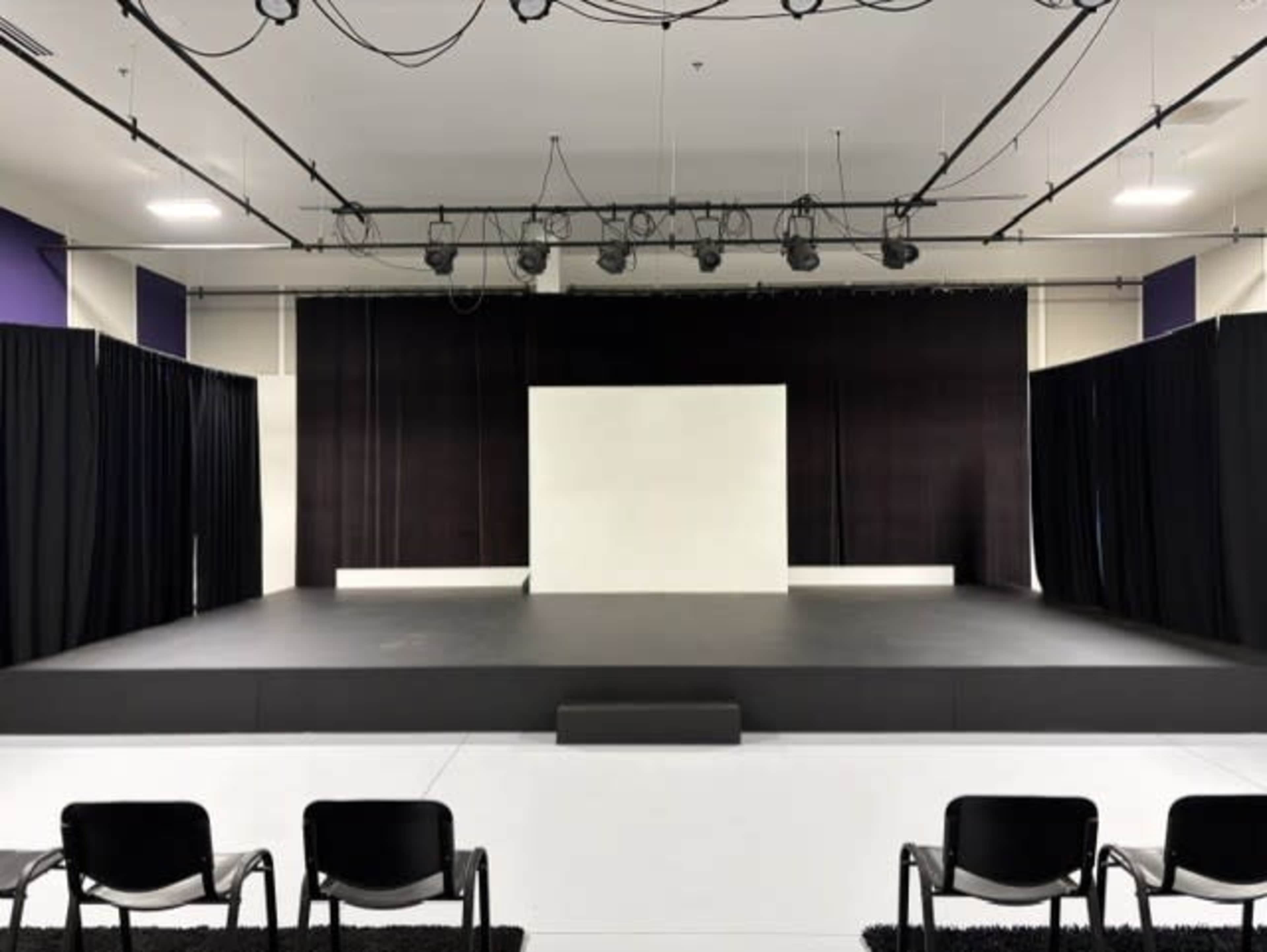The image shows a theater stage with a black curtain backdrop, a white screen in the center, and several black chairs arranged in front.