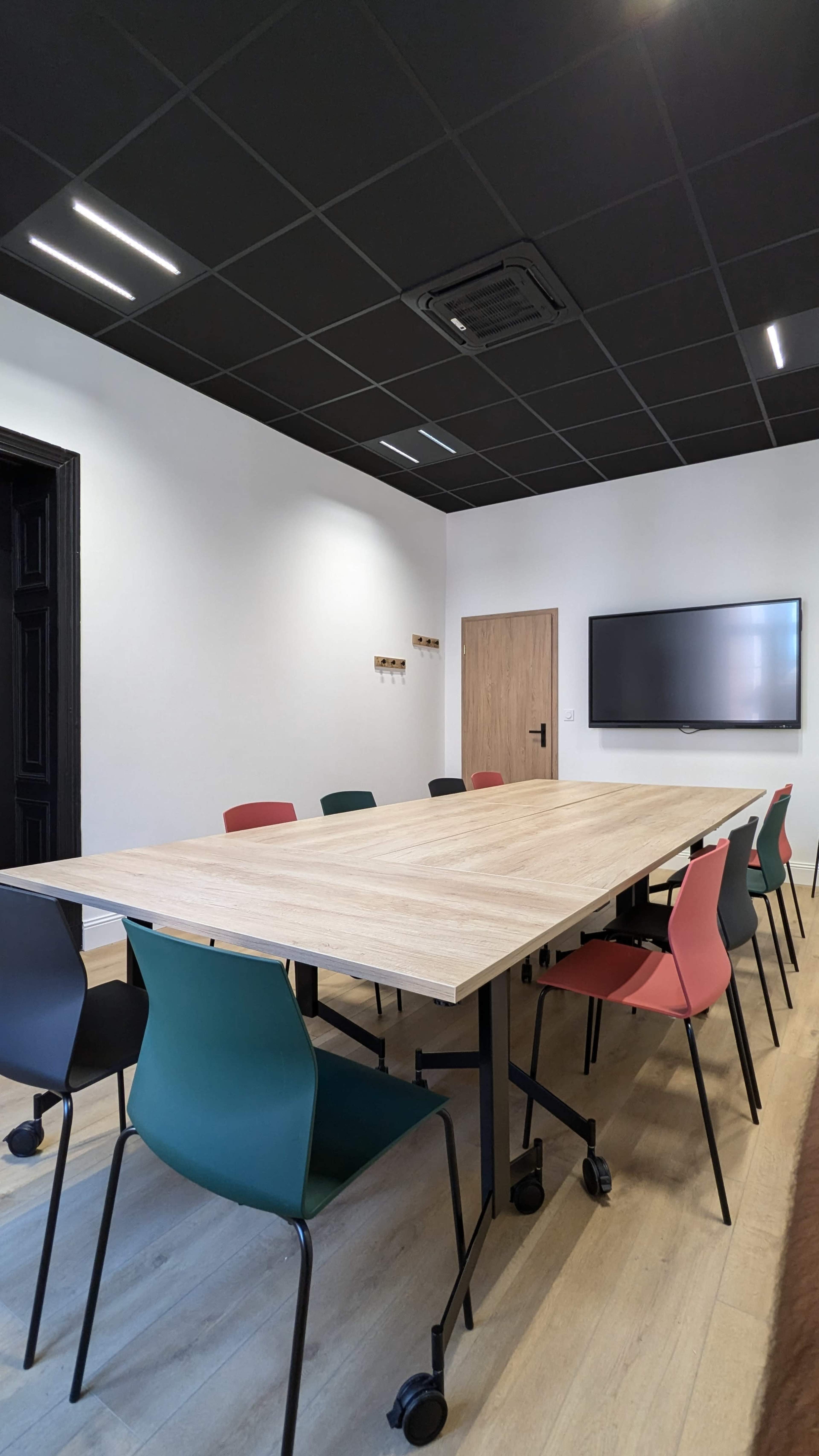 The image shows a modern conference room with a long wooden table surrounded by colorful chairs, a wall-mounted screen, and a minimalist design.